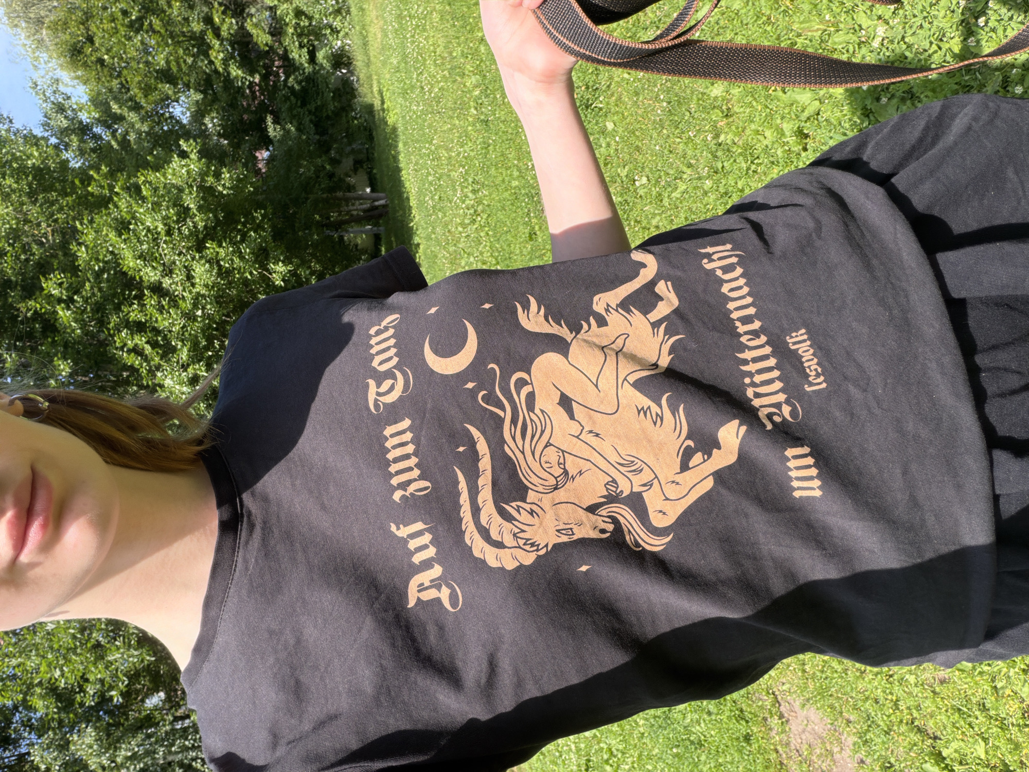 The image features a person wearing a black t-shirt with a golden graphic and text. The graphic depicts a mythical creature resembling a goat-like figure with horns and wings, intertwined with a human-like form with flowing hair. A crescent moon is also visible to the left of the creature.
The text on the t-shirt is in a Gothic-style font. On the left side, it reads "Auf und um". On the right side, it reads "um Mitternacht", with "Lesewolf" vertically positioned below "um Mitternacht".
The person is outdoors, with a background of green grass and trees, suggesting a park or natural setting. The lighting indicates it is daytime, likely sunny due to the shadows cast. The overall mood appears relaxed, given the casual attire and outdoor environment. The specific location cues are not immediately apparent beyond it being an outdoor, grassy area. The image seems to be a candid shot, possibly a selfie given the angle and the partial view of the person's face.