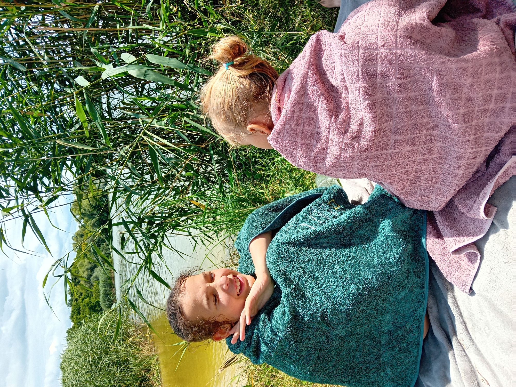 Two young girls, wrapped in towels, are sitting on a blanket by a body of water. The girl on the left, with brown hair pulled back, is wearing a teal towel and is leaning her head on her hands, with her eyes closed and a smile on her face. The girl on the right, with blonde hair in a bun, is wearing a pink towel and is facing away from the camera, looking towards the water. They are surrounded by tall green reeds and trees in the background. The water appears murky and yellow-green. The sky is partly cloudy. Based on the context, the location is likely a natural area with a lake or river, and is not specifically identifiable as being within the city of Chuhuiv, Ukraine.