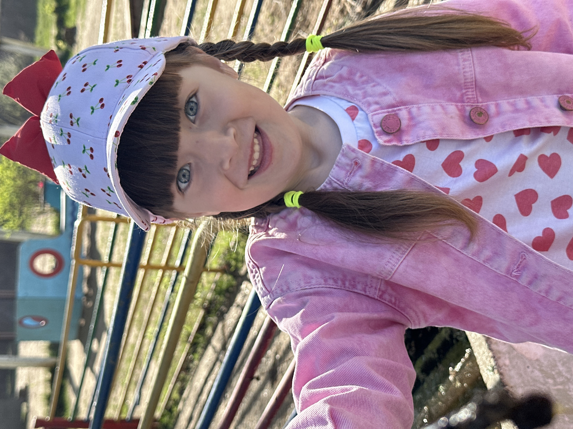A young girl is smiling at the camera, wearing a pink denim jacket over a white shirt with red hearts, a cherry-patterned hat with a red bow, and her brown hair is braided into pigtails with green elastic bands. She appears to be on a playground, as multi-colored bars are visible in the background. The photo was taken outdoors in daylight.