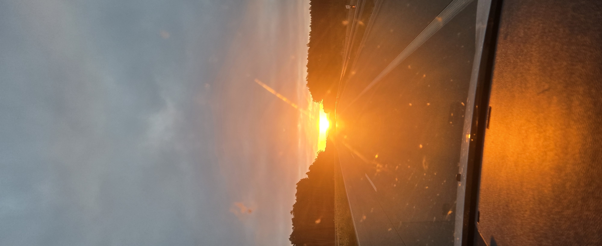 The image depicts a view from inside a vehicle, looking out at a sunset over a highway. The sun is low on the horizon, casting a vibrant orange and yellow glow across the sky and the road. The highway stretches into the distance, with trees lining both sides. The sky is filled with dramatic, dark clouds that contrast with the bright sunlight. The foreground shows the dashboard of the vehicle, illuminated by the setting sun. The location is identified as the city of Bożenkowo, Poland.