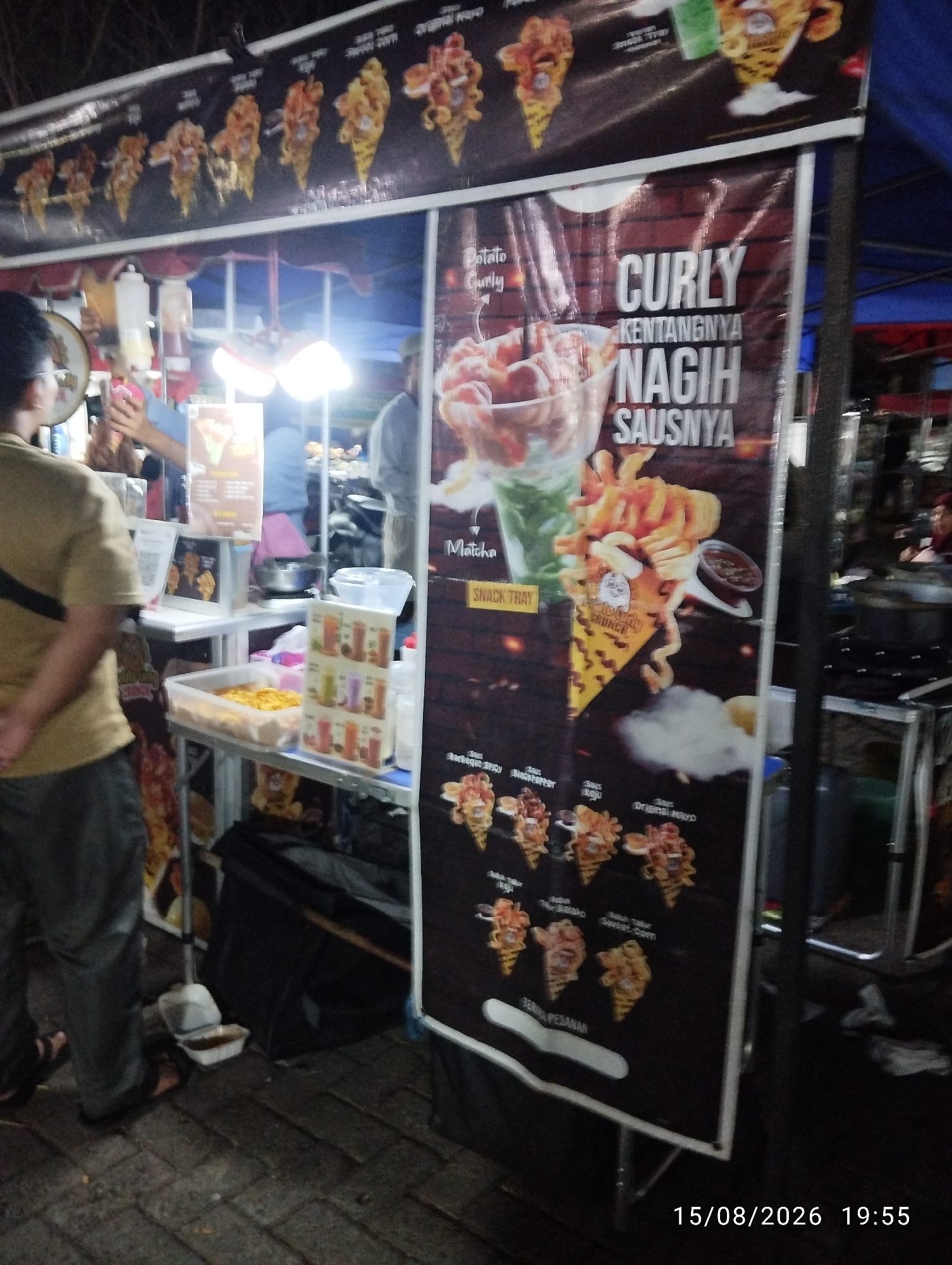 A night scene shows a street food stall with a large banner advertising curly fries. The banner features images of various curly fry cones with different sauces. A person is standing behind the counter, possibly an employee, preparing food. Another person, wearing a brown shirt and dark pants, is walking away from the stall. In the background, other stalls and people are visible, suggesting a busy market or street food area. The ground is paved with bricks, and there is a wooden cart with a display of drinks and snacks. The overall atmosphere is that of a lively night market.
