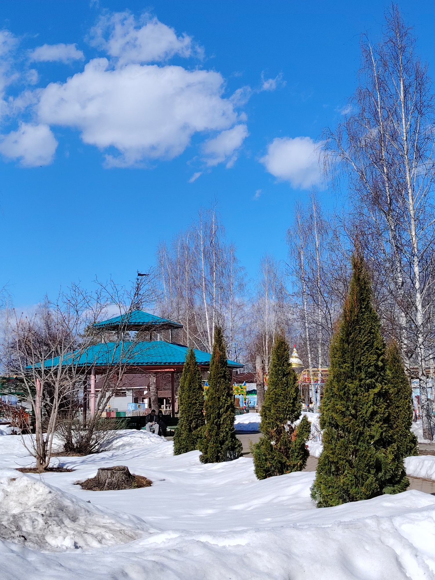 The image depicts a park scene in Chaikovsky, Russia during the late winter or early spring season. Patches of snow cover the ground. In the background, there is a gazebo with a distinctive turquoise, multi-tiered roof. Bare trees are interspersed with evergreen trees arranged in a line. People are visible sitting on a bench behind some bushes. The sky is clear blue, marked by scattered white clouds. A tree stump is visible in the snow in the foreground.