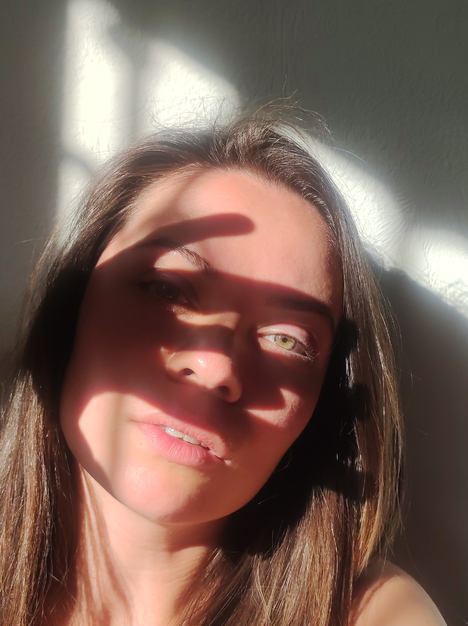 A close-up, high-angle selfie shows a young woman's face partially obscured by sunlight filtering through blinds. She has light brown hair and green eyes. The lighting creates strong shadows across her face, with a hand casting a shadow over her forehead and upper face. The background is a textured wall with more shadows. The image appears to be taken indoors, possibly in a bedroom or living area, with natural light. The woman is looking directly at the camera. There are no explicit indicators of location in Chaikovsky, Russia, within the image itself.