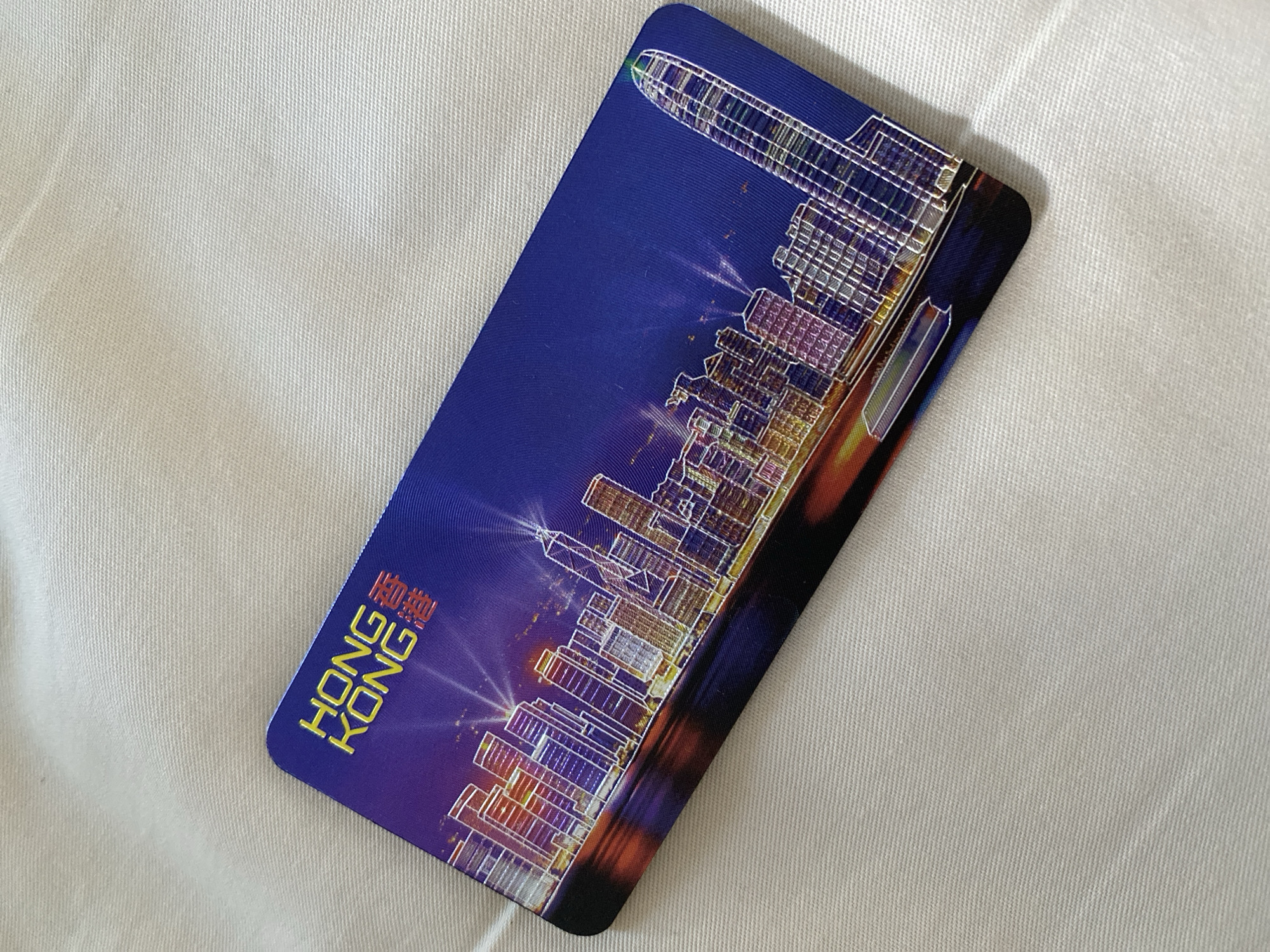 The image is a close-up, angled shot of a blue card featuring a nighttime cityscape. The card is placed on a textured white surface, likely fabric. The cityscape depicts a vibrant, illuminated skyline with numerous skyscrapers rendered in a stylized, outlined manner. Lights emanate from the buildings, and a body of water, reflecting the city lights with streaks of orange and yellow, is visible in the foreground. Text on the card reads "HONG KONG" in yellow lettering, accompanied by Chinese characters in red. A tall, prominent skyscraper with a pointed top is visible on the right side of the skyline. The card has rounded corners. There is no information in the image or provided context that places it in the city of North Kuta, Indonesia.