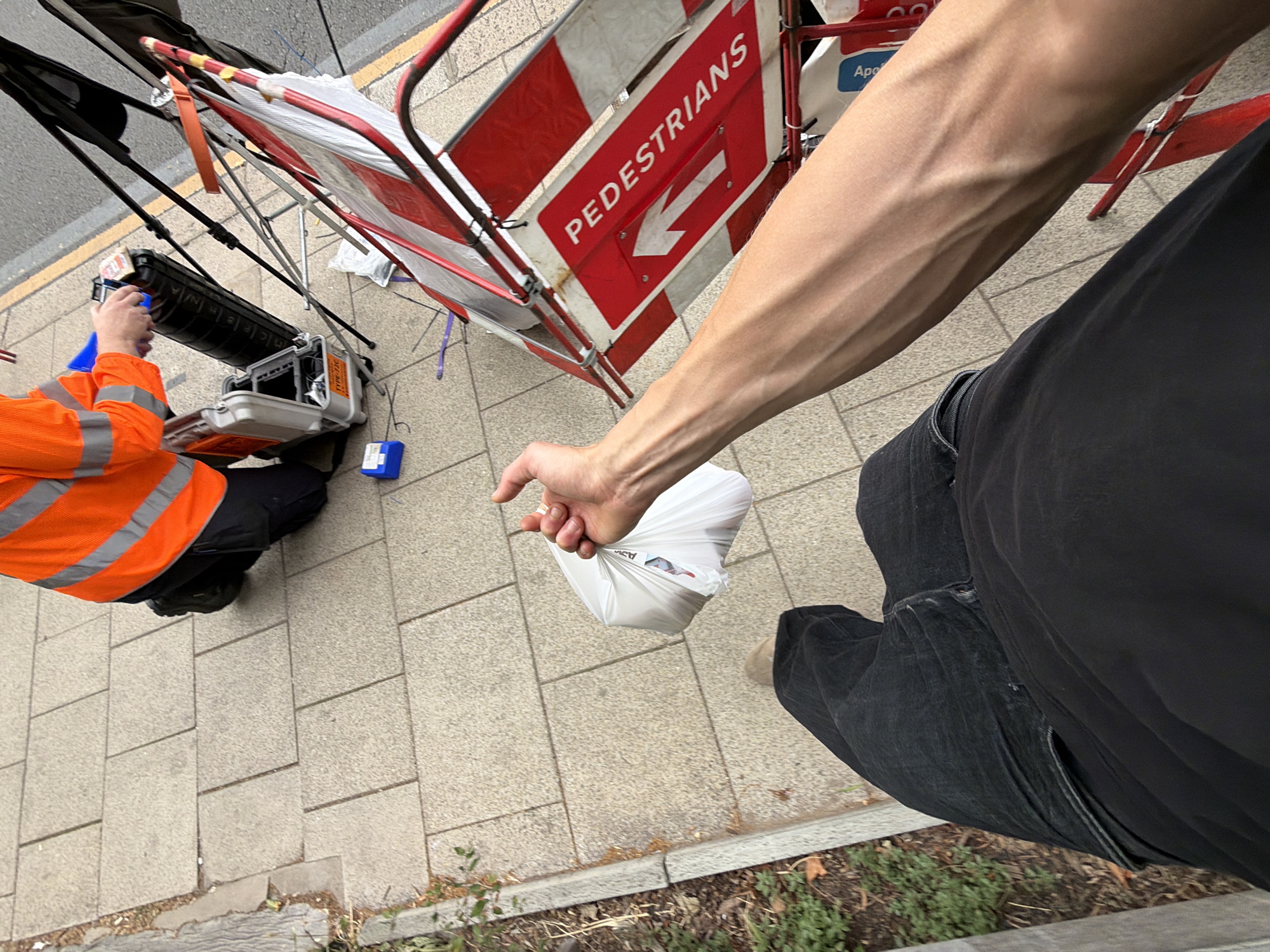 The image shows a person's arm and hand holding a white plastic bag, standing on a paved sidewalk next to a road closure. The person is wearing dark pants and their arm is prominent in the foreground, showing veins. Behind them, a construction barrier with red and white stripes is visible, along with a sign that reads "PEDESTRIANS" and has an arrow pointing left. A person wearing an orange high-visibility jacket is in the background, interacting with some equipment on the ground, possibly setting up or dismantling the barrier. The scene appears to be outdoors during the day. The paving stones suggest an urban environment, and the presence of a road closure and construction equipment points to some form of public works or maintenance. The overall mood is neutral, capturing a mundane moment during what seems to be work being done on a street. There is no clear indication of weather conditions, but the lighting suggests it is daytime.