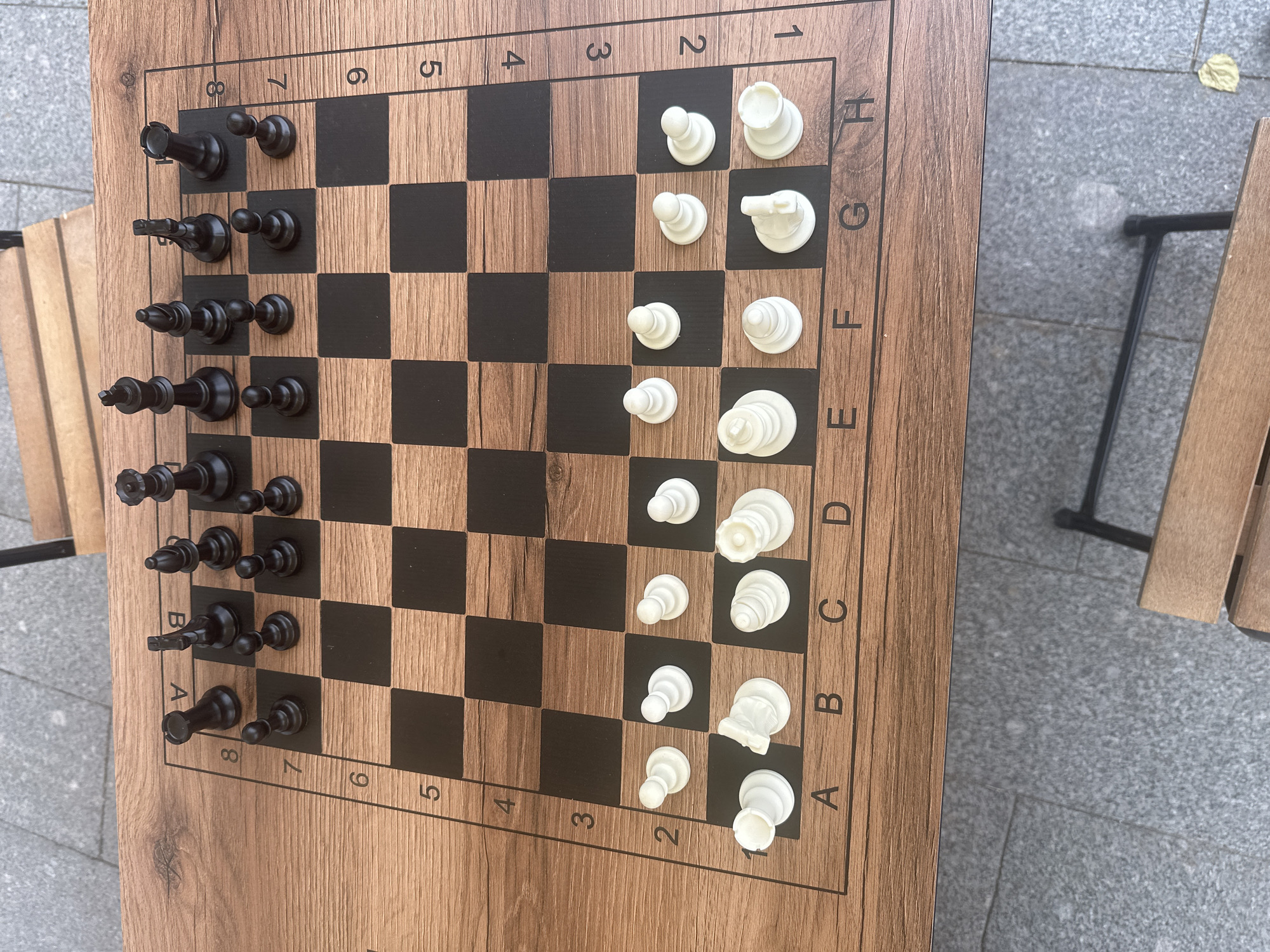 An overhead, high-angle shot shows a wooden chess board with a wood grain pattern and black squares. The chess pieces, black on one side and white on the other, are set up in their starting positions. The black pieces are on ranks 7 and 8, with pawns on rank 7 and the remaining pieces on rank 8. The white pieces are on ranks 1 and 2, with pawns on rank 2 and the remaining pieces on rank 1. The chess board is placed on a rough, grey, paved surface that appears to be outdoors. The background is blurred, but hints of a wooden bench and metal chair legs suggest an outdoor seating area. The overall scene is static and peaceful. No people are visible in the image, and there are no discernable landmarks or clues that would indicate the location as being in Moscow, Russia.