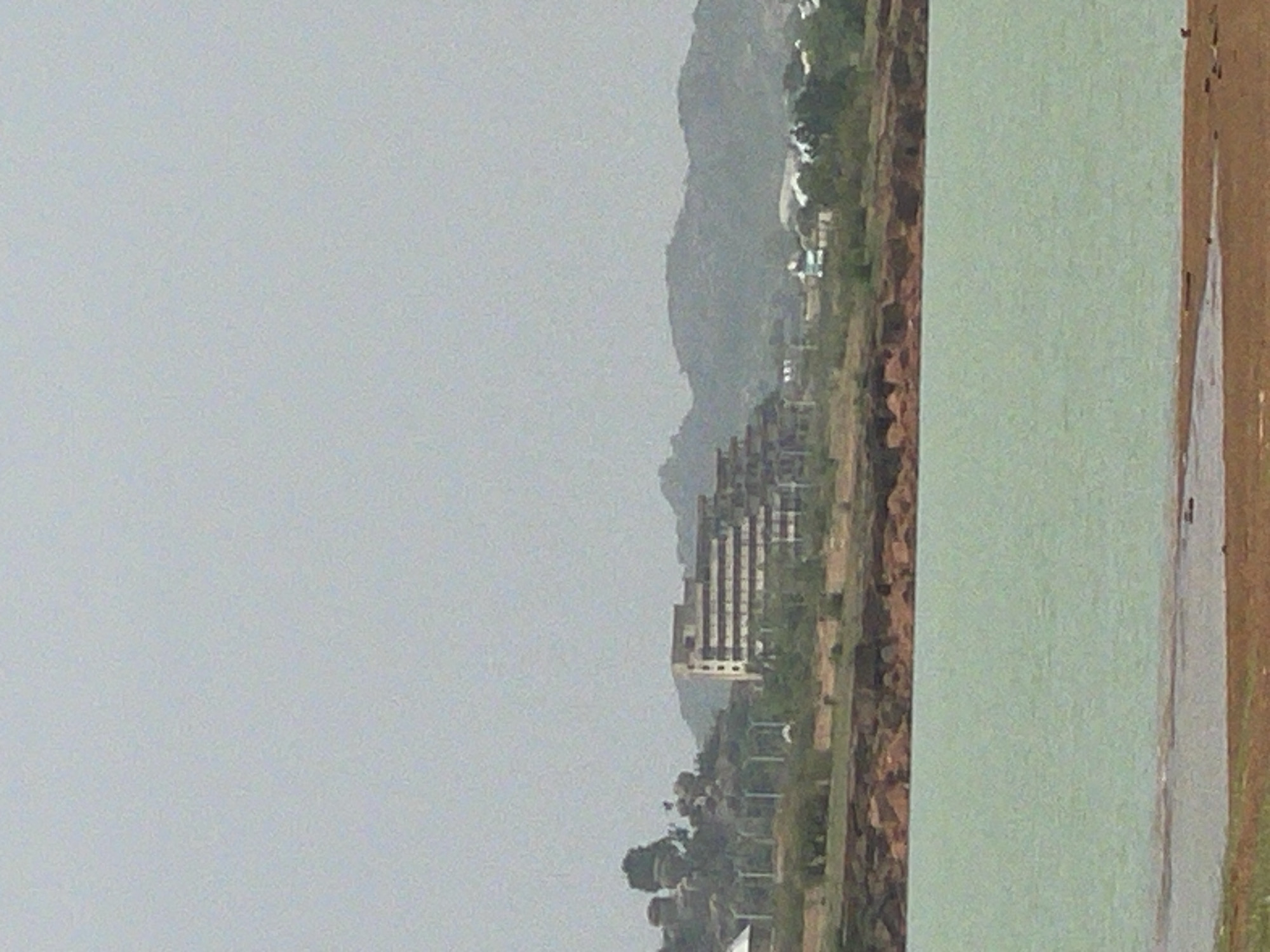 The image shows a landscape of Nabatum, Nigeria on an overcast day. A body of water is on the right side of the frame, bordered by a brown shoreline. The central part of the image contains a stretch of land with sparse vegetation, including trees and low-lying plants. A high-rise building stands out against the backdrop of distant hills, suggesting the presence of urban or developed areas within the natural scenery. The sky is uniformly gray, indicating a cloudy or foggy weather condition.