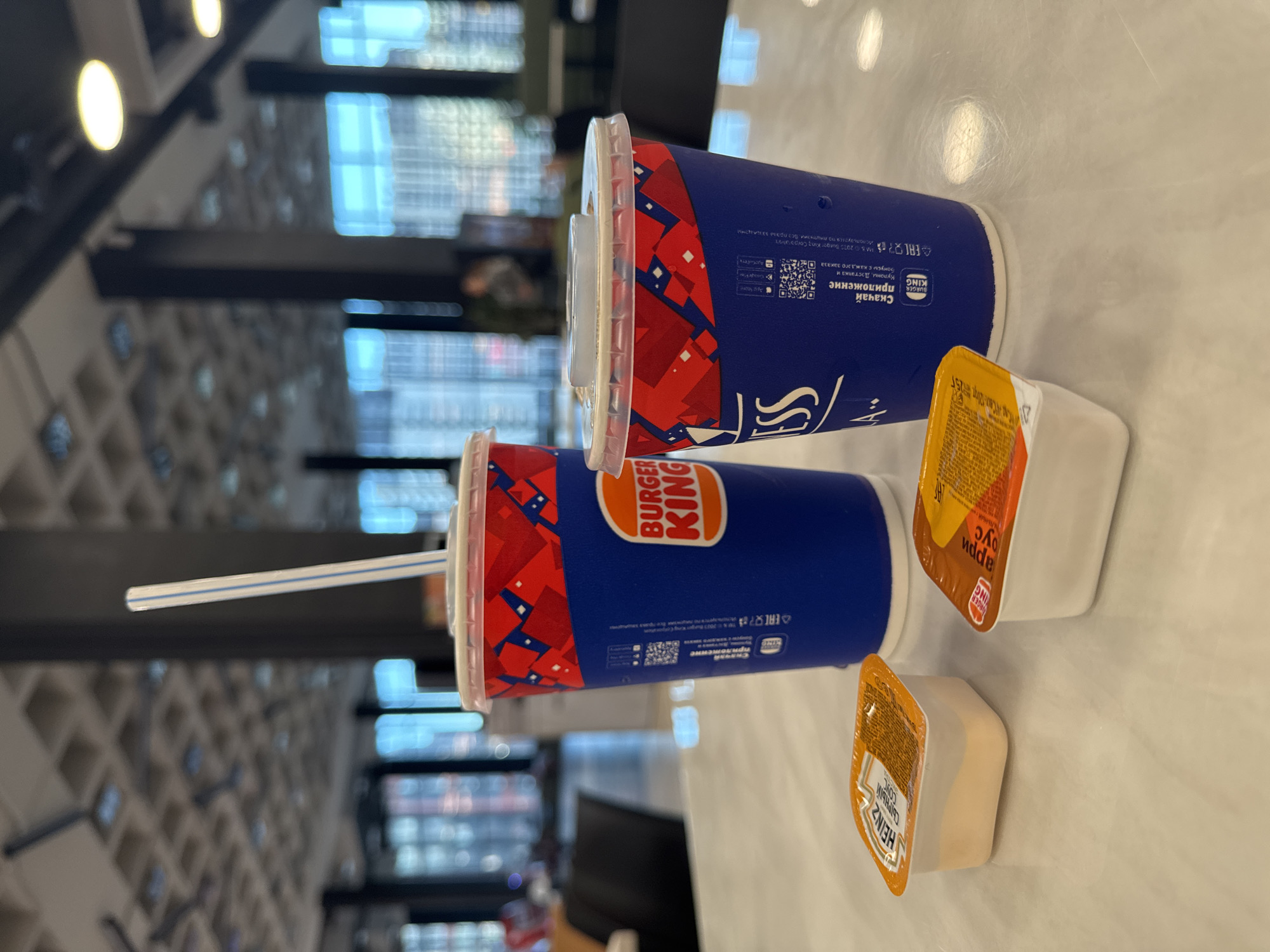 The image shows a close-up of two Burger King drinks and two dipping sauce containers on a table in what appears to be a Burger King restaurant. The drinks are in blue cups with red and white Burger King branding. One cup has a white and blue striped straw sticking out. The dipping sauce containers are small, square, and appear to be cheese sauce and apple sauce, based on the labels. The background is blurred but suggests an indoor setting with tables and chairs, possibly a food court or a restaurant dining area. The lighting is bright, indicating overhead lights. There are no people clearly visible in the image. The overall scene is a typical fast-food meal setup.