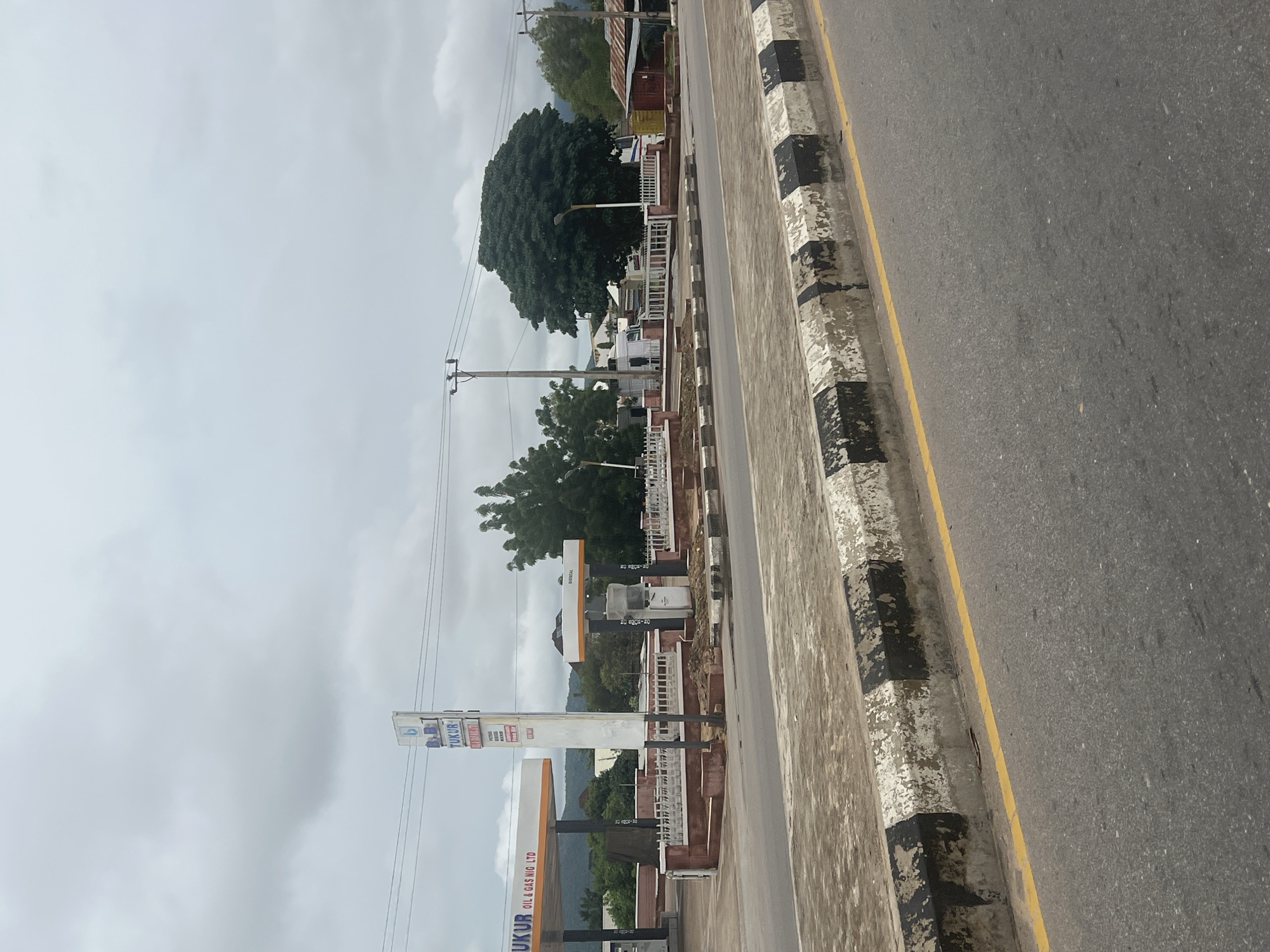 The image captures a view of a gas station in Jalingo, Nigeria, on an overcast day. A tall white sign with blue lettering, reading "B.B. TUKUR OIL & GAS NIG. LTD," stands prominently near the entrance of the station. To the left, a white and orange gas canopy covers several fuel pumps. To the right, another gas pump island, also with a white and orange canopy, is visible, and it is labeled "DIESEL." A white dispenser is positioned at this island. The gas station area is demarcated by a low reddish-brown wall and a white picket fence. Beyond the gas station, a line of trees forms a backdrop, with some buildings and power lines visible. In the foreground, a road is present with a yellow line marking its edge, and a concrete curb painted with alternating black and white stripes. The ground appears to be paved, with some parts of the curb showing wear and tear. No people are visible in the scene.
