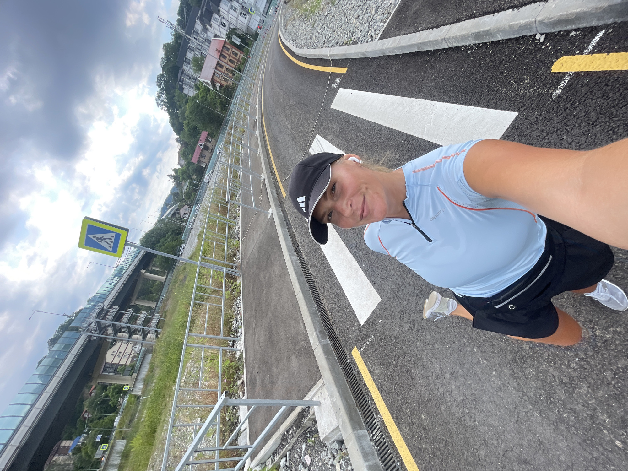 The image shows a woman in athletic wear, including a light blue t-shirt, black shorts, a black baseball cap, and white sneakers. She is wearing wireless earbuds and a small black fanny pack. The woman is smiling at the camera, suggesting a positive emotion, possibly enjoyment of her activity or the scenery.
She appears to be running or walking on a paved road with yellow and white markings, indicating a pedestrian crossing. The road curves to the right in the foreground. In the background, there are buildings on a hill, a large pedestrian overpass or bridge with a glass enclosure, and lush green trees. A yellow pedestrian crossing sign is visible on the left side of the image. The sky is cloudy, suggesting overcast weather. The overall setting appears to be an urban or semi-urban area with significant infrastructure, likely a park or recreational path near a road.
The image is taken from a low angle, possibly a selfie taken by the woman herself while in motion. The orientation of the image is tilted, with the woman and the road appearing at an angle. The text "CRAFT" is visible on the woman's t-shirt. The presence of the pedestrian overpass and the well-maintained road suggest this could be a developed area within Sochi, Russia, as indicated by the location context. The time of day is likely daytime, given the lighting conditions, though the cloudy sky makes it difficult to determine precisely.