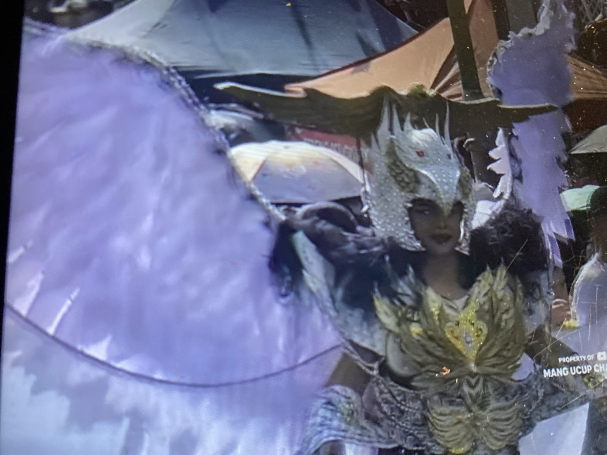 The image depicts a fragmented and distorted view of what appears to be a performance or elaborate costume display. A large, purple, fabric-like structure dominates the top portion of the frame, with textured elements suggesting feathers or scales. Beneath this, several figures or objects are visible, partially obscured and disoriented. One figure wears a headdress or mask adorned with gold and green, featuring feathery or leafy elements. Other areas show glimpses of shimmering, scale-like textures in silver and grey, as well as darker, possibly fabric or leather materials. The scene is overlaid with a watermark indicating "PROPERTY OF MANG UCUP CHA," suggesting a personal recording or documentation from Ketileng, Indonesia. The overall impression is of a vibrant, possibly ceremonial or artistic display, captured with low fidelity and a skewed perspective.