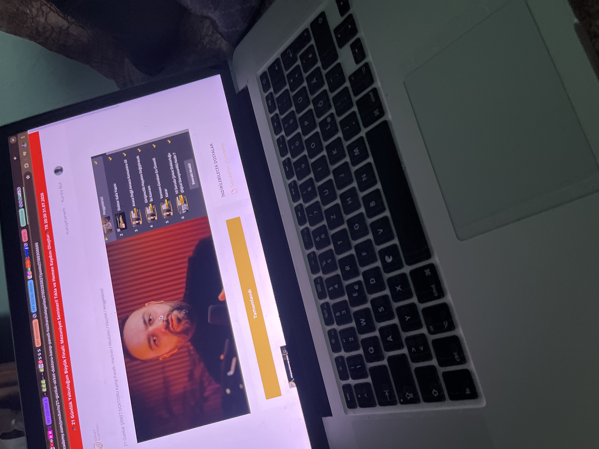 A laptop screen displays a webpage titled "21 Günlük Yolculuğun Büyük Finali: Mezuniyet Semineri' Tıkla ve Hemen Kaydını Oluştur". The page features a video of a man speaking, with a list of course modules on the right side. Below the video is a yellow button labeled "Tamamlandı". The laptop's keyboard is visible in the foreground. The image is taken indoors, likely in a residential setting, given the lighting and background which appears to be a fabric surface. There is no visual information within the image to indicate the city of Pet Mogili, Bulgaria.