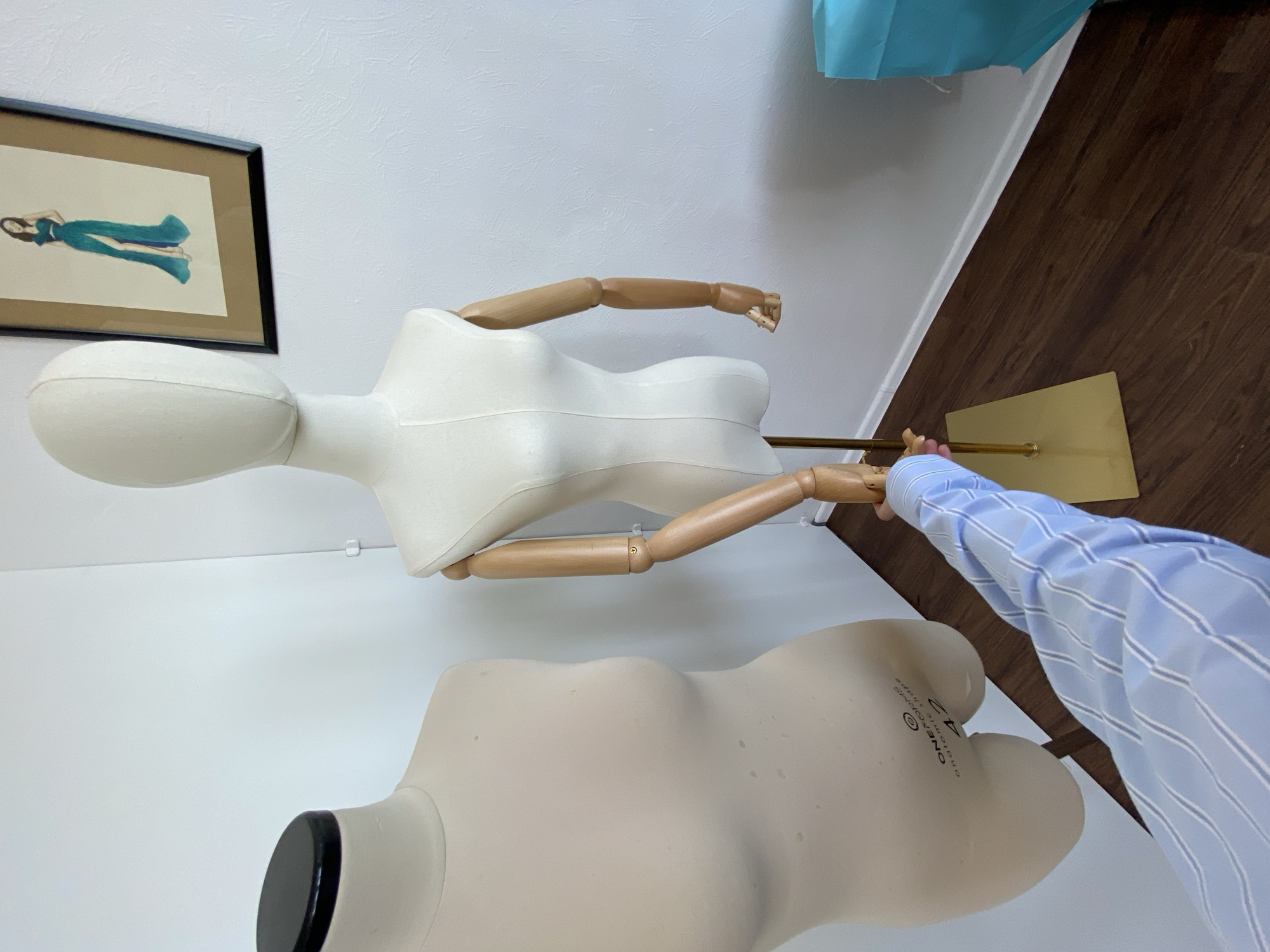 The image shows a room with a white wall and a wooden floor. There are two mannequins; one is white with wooden arms, and the other is a light beige color. A person in a blue striped shirt is seen adjusting the arm of the white mannequin. On the wall, there is a framed picture. A piece of blue fabric is visible in the upper part of the frame. Based on the limited visual information, it is not possible to confirm the location of the image as Izhevsk, Russia.