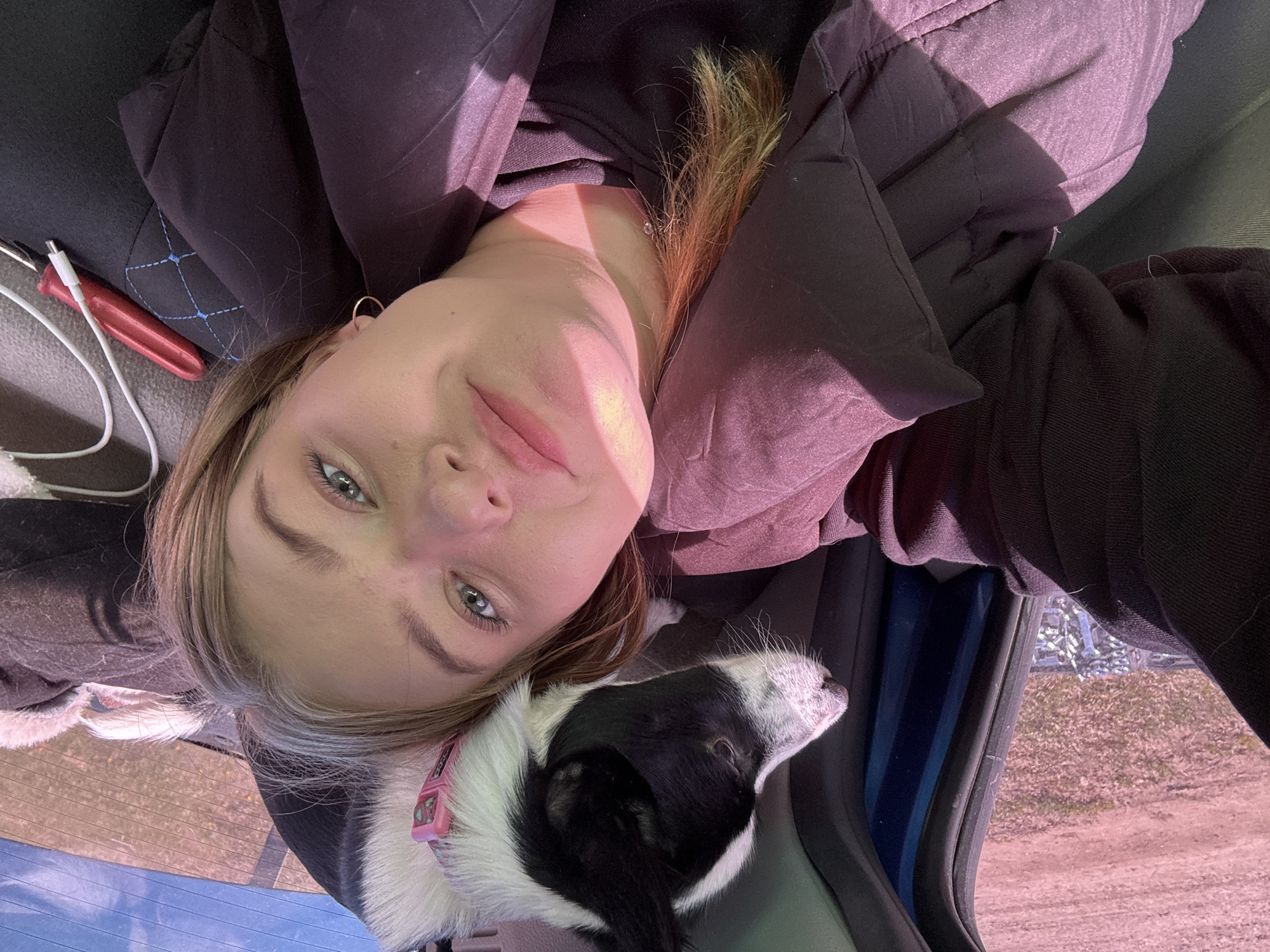 A young woman is in a car with a black and white dog. The woman is wearing a dark jacket and has blond hair. She is looking at the camera. The dog is wearing a pink collar. The car is parked on a dirt road. The location is in Узяр, Russia.