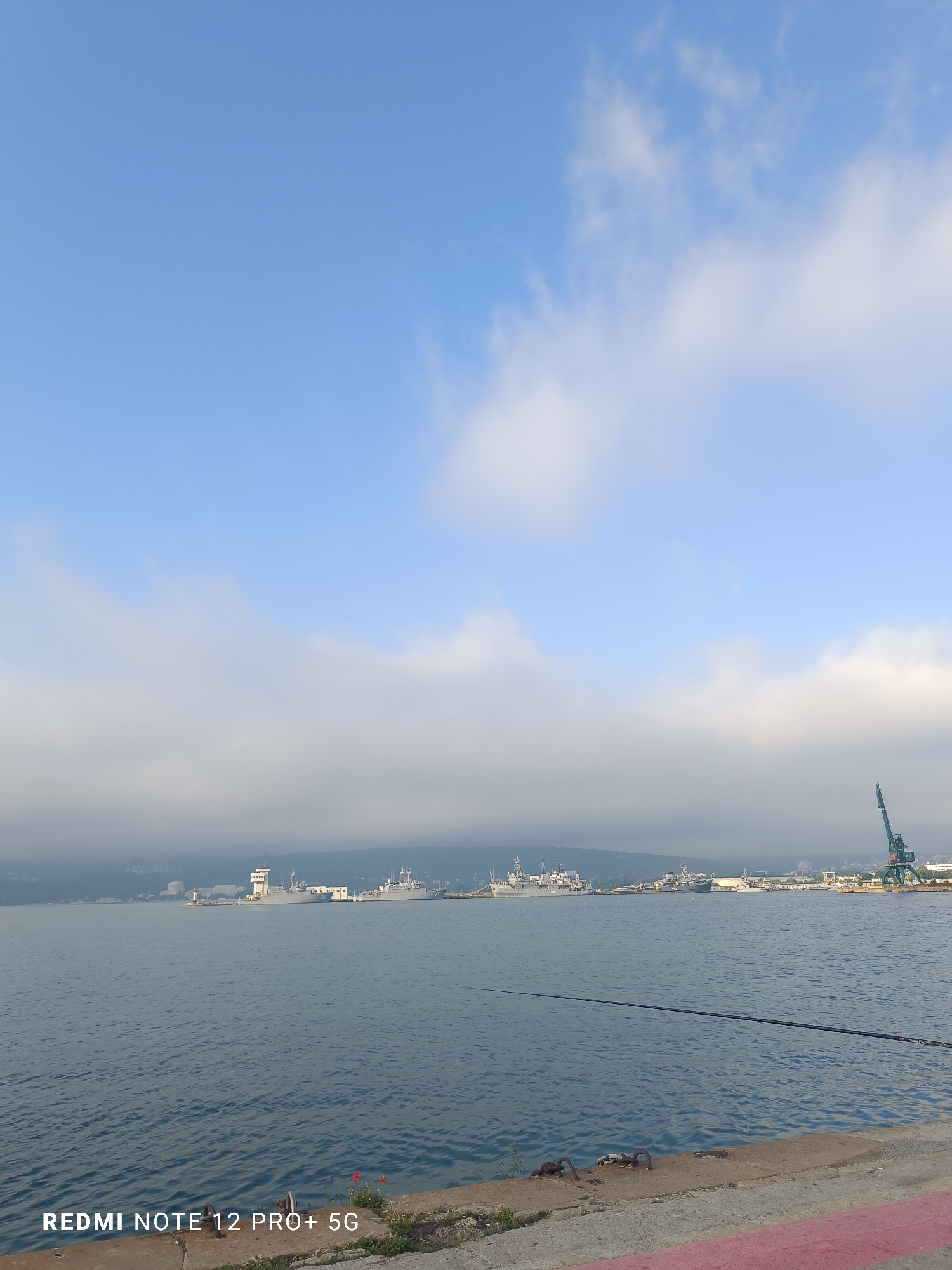 The image captures a wide view of a calm harbor or port area, likely in Varna, Bulgaria, under a partially cloudy sky. In the foreground, a concrete pier or embankment is visible, extending across the bottom of the frame. Several metal mooring rings are embedded in the concrete, and a red painted line marks the edge on the right. A fishing rod extends diagonally from the right edge into the water, suggesting an ongoing fishing activity.
The midground features a large expanse of still, blue-grey water with subtle ripples. Along the distant shoreline, several large ships, possibly naval vessels or cargo carriers, are docked. A prominent white building with a tower, resembling a control tower or lighthouse, stands out among the structures on the far shore. Hills or mountains, partially obscured by low-lying mist or clouds, form the backdrop. On the far right of the distant shore, a tall green industrial crane is visible, indicating active port operations.
The sky above is bright blue with scattered white, wispy clouds, particularly in the upper left. A distinct band of white, hazy clouds or mist stretches across the middle of the sky, blending with the distant land and obscuring the upper parts of the hills. The lighting is soft, suggesting either early morning or late afternoon.
In the bottom left corner of the image, the text "REDMI NOTE 12 PRO+ 5G" is clearly visible in white capital letters. The overall scene conveys a serene atmosphere, combining natural elements with industrial activity.