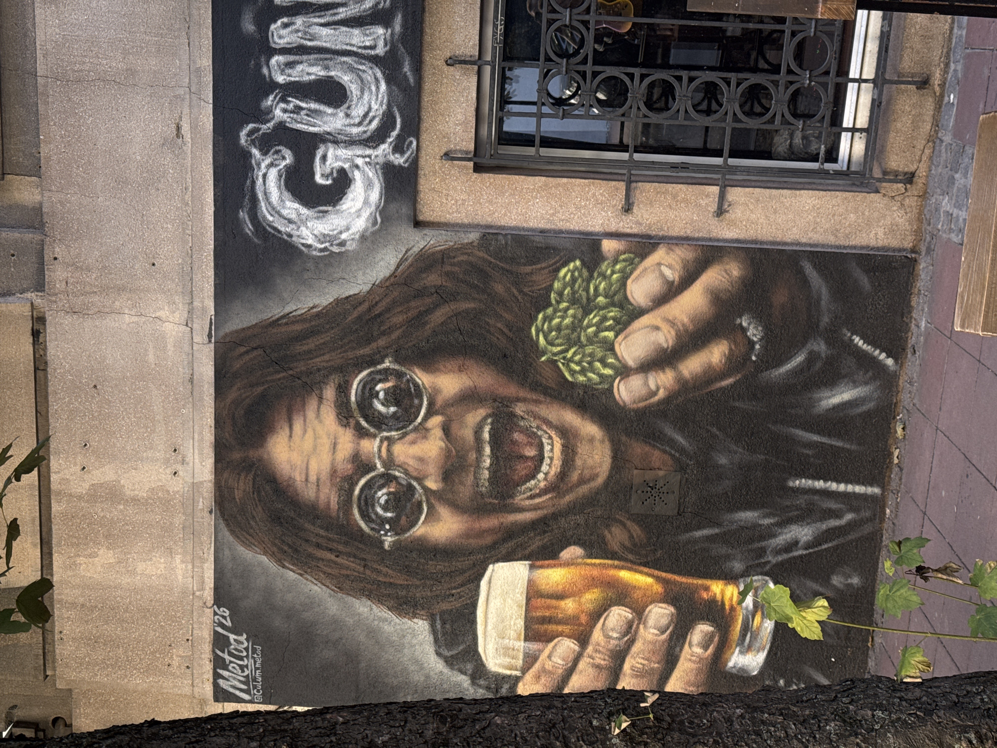 This is a mural painted on the exterior wall of a building in Belgrade, Serbia. The mural depicts a person, likely a man, with long brown hair and round glasses, holding a glass of beer in one hand and a cluster of hops in the other. The person has their mouth open in a shouting or singing expression. Above the mural, to the right, the word "GUM" is spray-painted in white. To the left of the mural, near the top, the signature "Metod 26" and a social media handle "@Culom.metod" are visible. The mural is cracked in several places, indicating age or damage. A tree trunk is positioned to the left of the mural, and a window with a decorative metal grate is to the right. The ground in front of the mural is paved with reddish-brown tiles.