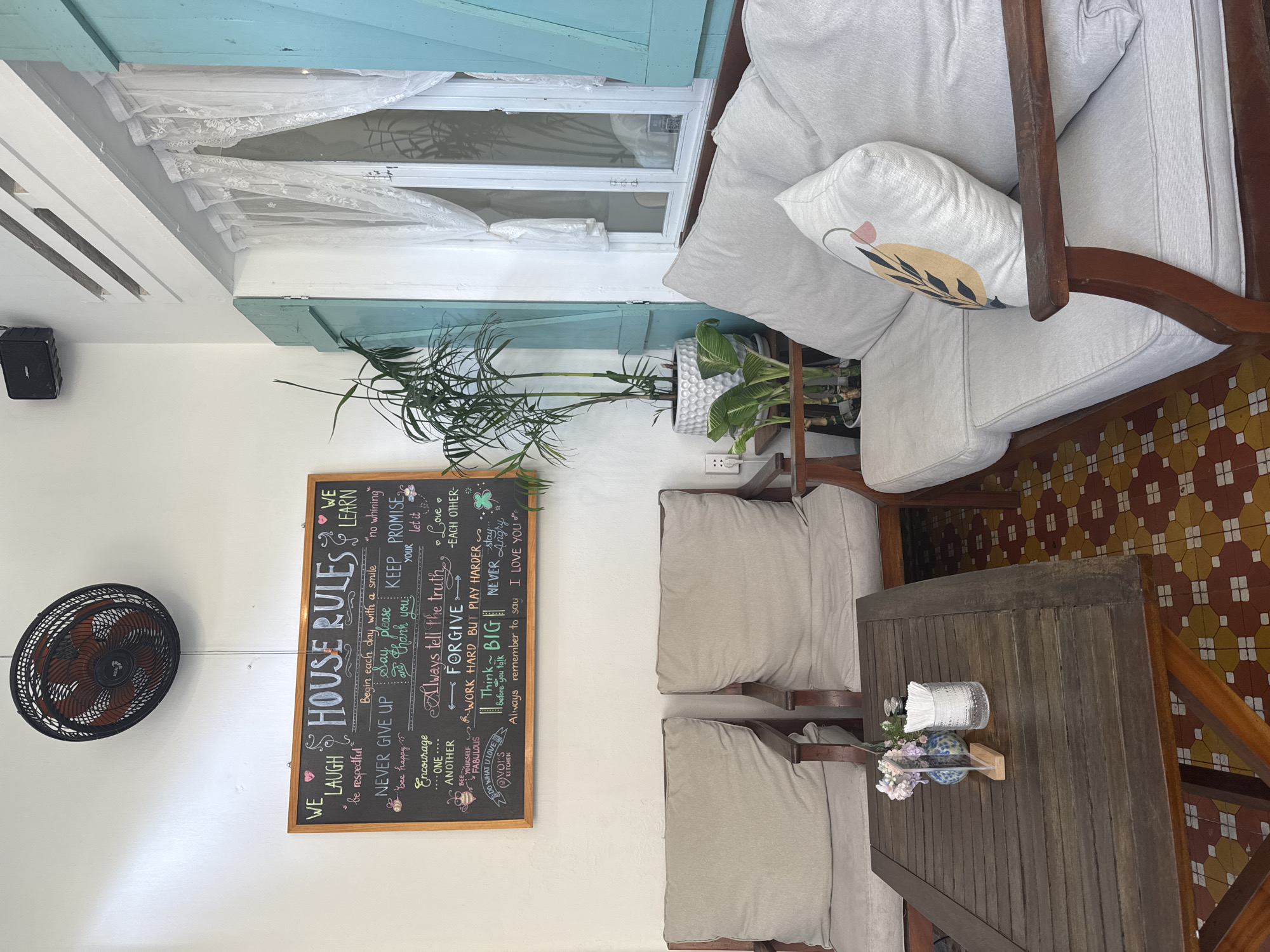 The image depicts an interior space, likely a room or café, in Đà Nẵng, Vietnam, based on the style and décor. A framed chalkboard labeled "HOUSE RULES" is prominently displayed against a white wall, featuring a list of positive affirmations and guidelines written in chalk. To the left of the chalkboard, a black wall-mounted fan is visible. Above the chalkboard, there are plants in pots, and a window covered with lace curtains and blue shutters. Furniture includes a wooden table with place settings, napkins, and a small floral arrangement. Two cushioned chairs flank the table, and a cushioned loveseat is visible in the upper right corner of the frame. The floor is tiled in a patterned mosaic of red, yellow, and white. The overall atmosphere is casual and inviting.