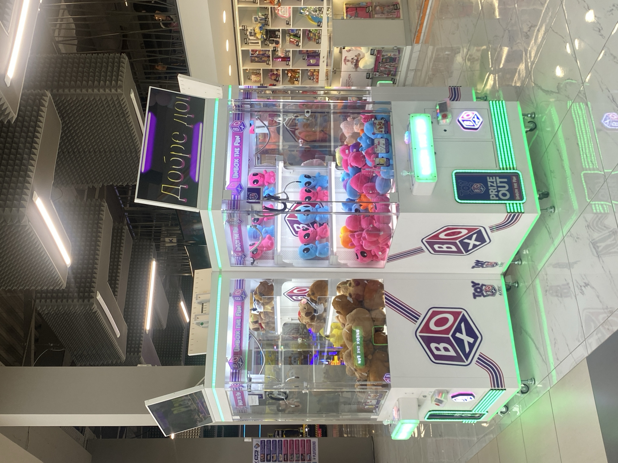The image shows two large, white claw machine arcade games in what appears to be a shopping mall. The machines are filled with plush toys, including teddy bears and alien-like figures in various colors like pink, blue, and orange. The machines have illuminated tops and sides, with colorful branding that includes the text "BO" inside a hexagon and "UNBOX THE FUN". One machine has a sign that reads "PRIZE OUT UNBOX THE FUN". In the background, a shelf-lined store is visible, displaying various items. The floor is tiled with a light marble pattern. The scene is indoors, under artificial lighting, and the overall atmosphere suggests a commercial entertainment or retail environment. The sign at the top of one machine displays text in a Cyrillic script, which translates to "Welcome". The presence of this phrase in Cyrillic, combined with the architectural style and typical shopping mall layout, strongly indicates that this location is in Bulgaria. Specifically, considering the prevalence of such entertainment options, a likely location for this image to have been taken is within the central atrium or a designated entertainment zone of a prominent shopping mall in Burgas, Bulgaria, such as the Galleria Burgas. No individuals are clearly visible in the foreground.