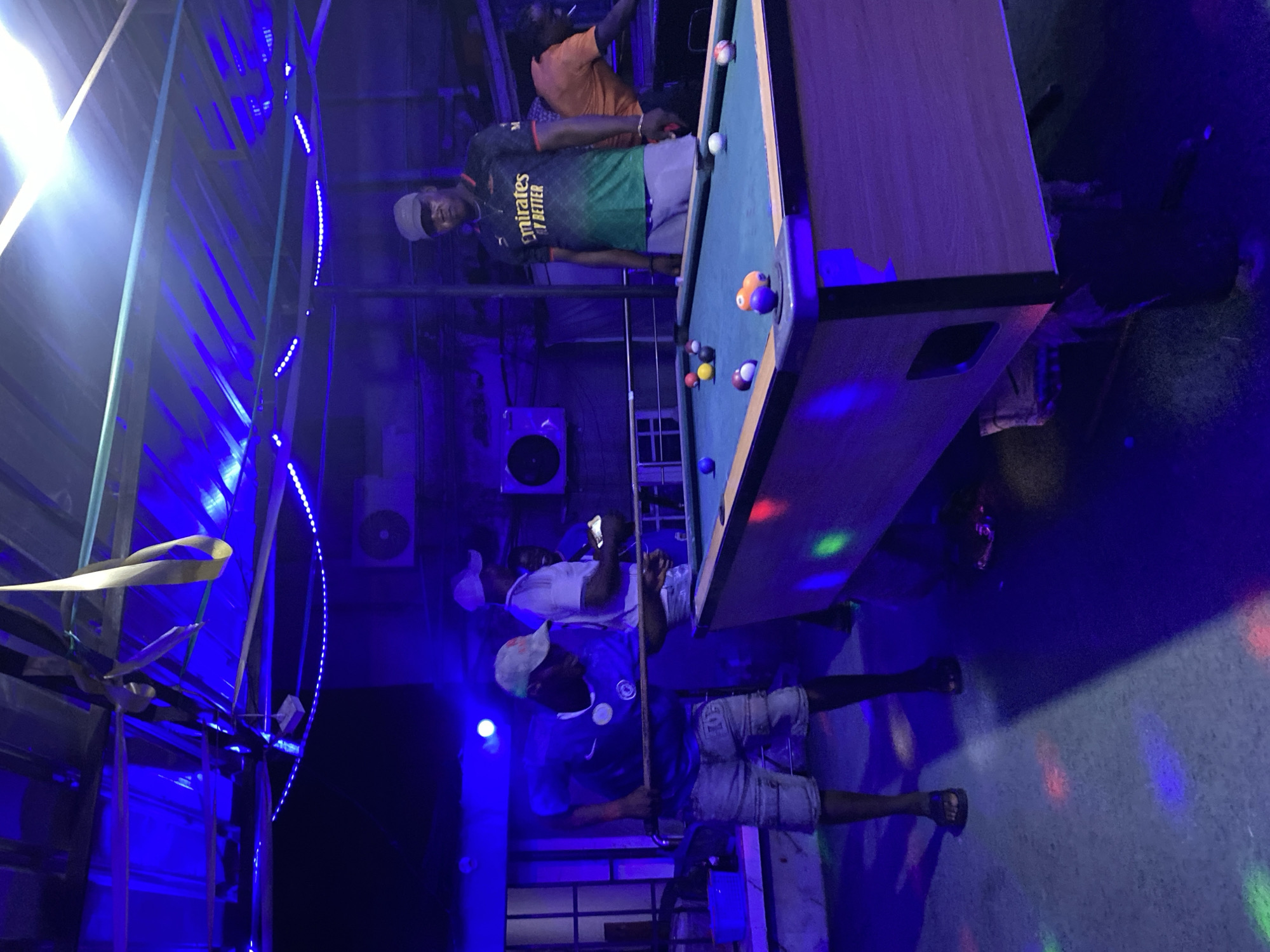 Ado, Nigeria: An indoor scene reveals two men playing pool under blue lighting. One man, wearing an "Emirates Fly Better" shirt, stands beside the pool table. Another man is holding a pool cue, with a phone in his left hand. An adjacent structure is illuminated with blue LED string lights. Another man is standing behind the pool table.