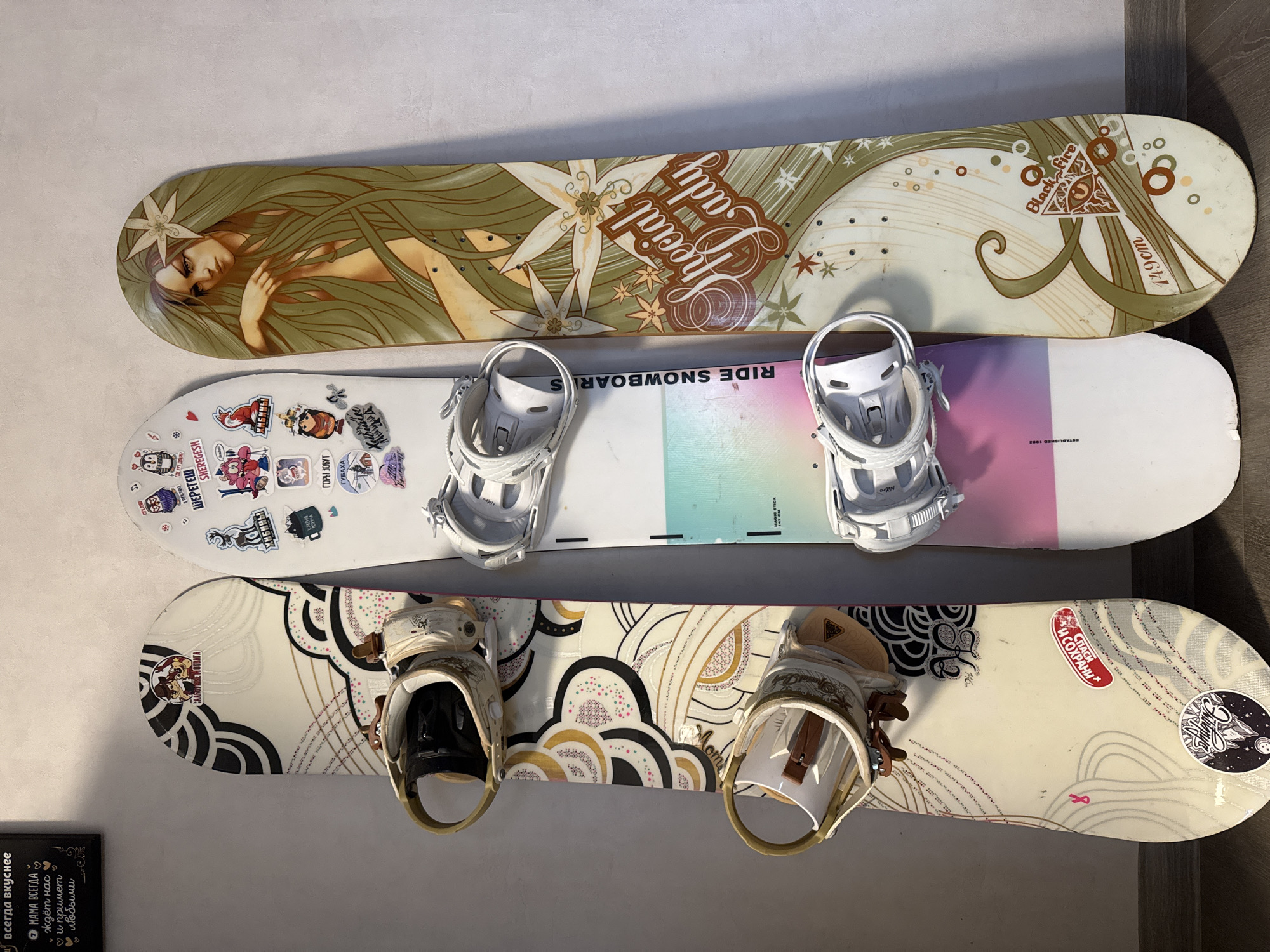 Three snowboards with bindings are stacked vertically against a light-colored wall, likely indoors, with a darker floor visible to the right. The snowboards appear to be personal equipment, suggesting storage in a home or gear room in Chaikovsky, Russia.
The top snowboard features an elaborate graphic of a woman with long, flowing green hair and a flower, with the stylized text "Special Lady" prominently displayed. Towards the tail, a triangular symbol with an eye and the text "Black Fire" and "149cm" are visible. Its bindings are white.
The middle snowboard is predominantly white with a vibrant, pastel gradient of pink, purple, and light blue towards the tail. It is covered with various stickers, some featuring cartoon characters and others bearing text in Cyrillic, including "SHEREGESH," "ХИБИНЫ" (Khibiny), and "ГУБАХА" (Gubakha), which are names of Russian ski resorts or regions. The brand "RIDE SNOWBOARDS" is visible on the base, and "Nitro" is seen on one of the white bindings. Faint text also reads "MAGIC STICK 147 CM" and "ESTABLISHED 1992".
The bottom snowboard has a cream-colored base adorned with a black and gold abstract pattern resembling arches or waves, interspersed with small, colorful dots. A sticker near the nose reads "СЛАБОУМИЕ И ОТВАГА" (Weak-mindedness and Courage). Another sticker on the tail says "СПАСИ И СОХРАНИ" (Save and Protect), and a small pink ribbon symbol is also present. Its bindings are cream or gold-colored.
In the bottom left corner, part of a dark sign or board is visible, displaying Cyrillic text: "Всегда вкуснее" (Always tastier) and "МАМА ВСЕГДА ждёт нас ♡ и примет • любыми ♡" (Mom always waits for us and will accept us any way). No activity is taking place, and the time of day or weather cannot be determined.