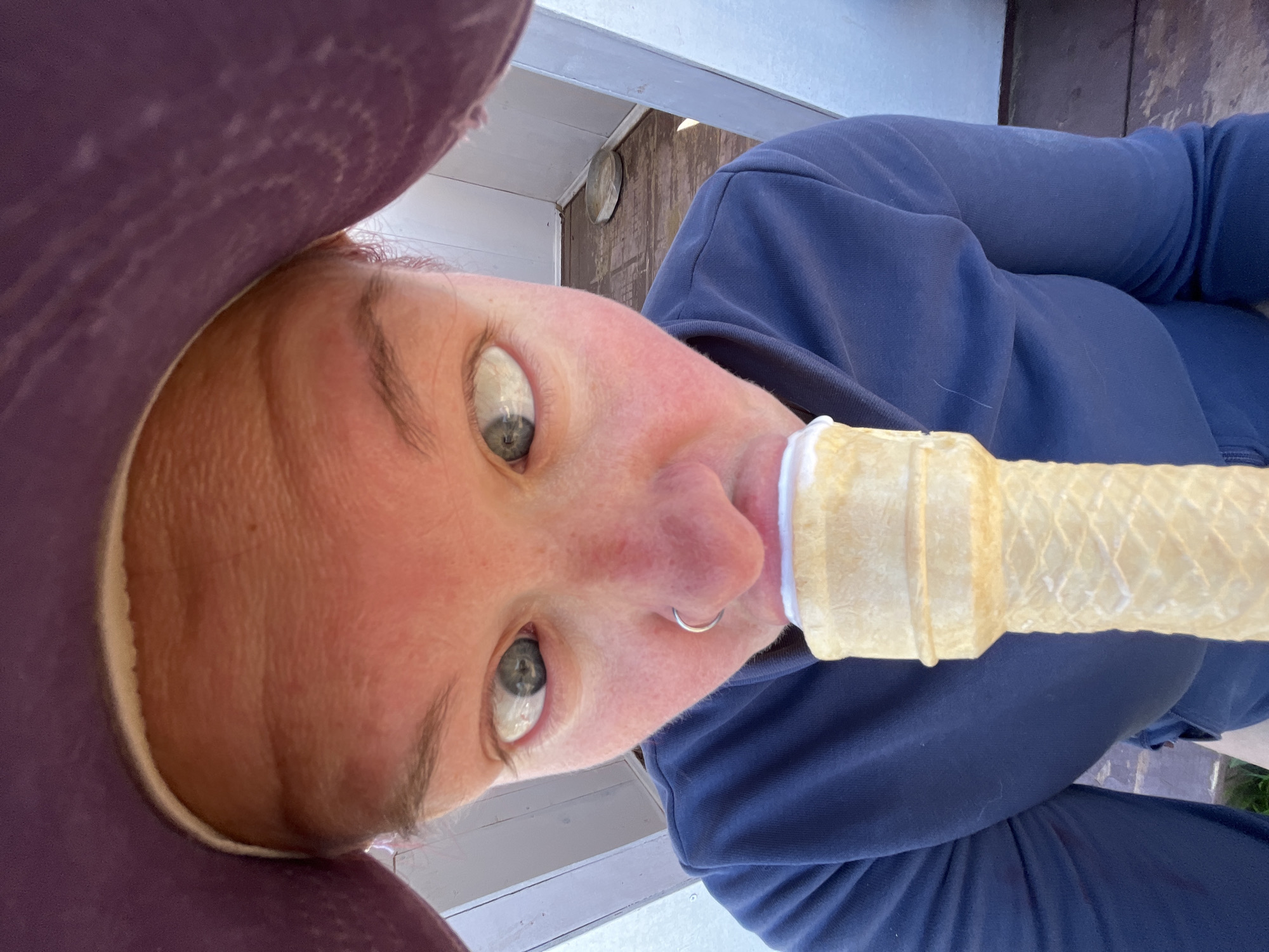 A close-up, low-angle shot depicts a person with pale skin, light-colored eyes, and a nose piercing. They are wearing a dark blue sweatshirt and a burgundy hat. In their mouth, they hold a rolled-up piece of yellowish material, resembling a large, hollow edible tube. The background shows a partially visible wooden structure with white painted trim. The location appears to be outdoors, possibly on a porch or deck. There is no indication that the image was taken in Chaikovsky, Russia.