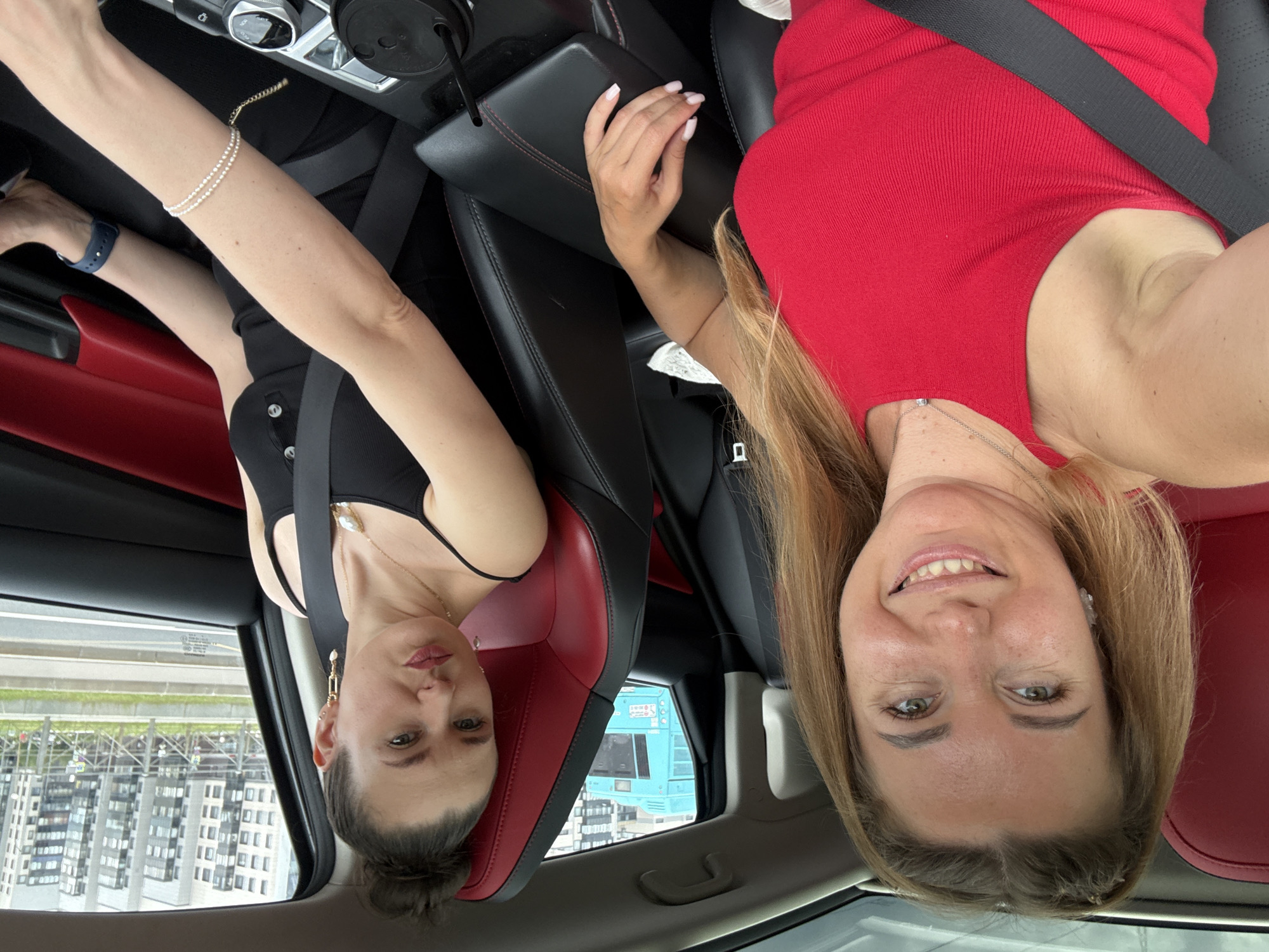 Two women are in the front seats of a car, looking at the camera. The woman in the foreground, on the left, is smiling and has blonde hair pulled back into a ponytail. She is wearing a red tank top and a silver necklace. The woman in the background, on the right, has her hair in a bun and is wearing a black tank top and gold earrings. She is looking at the camera with her lips pursed. They are both wearing seatbelts. The car's interior has red and black leather seats. Outside the car, buildings and a blue bus are visible. The setting appears to be an urban environment.