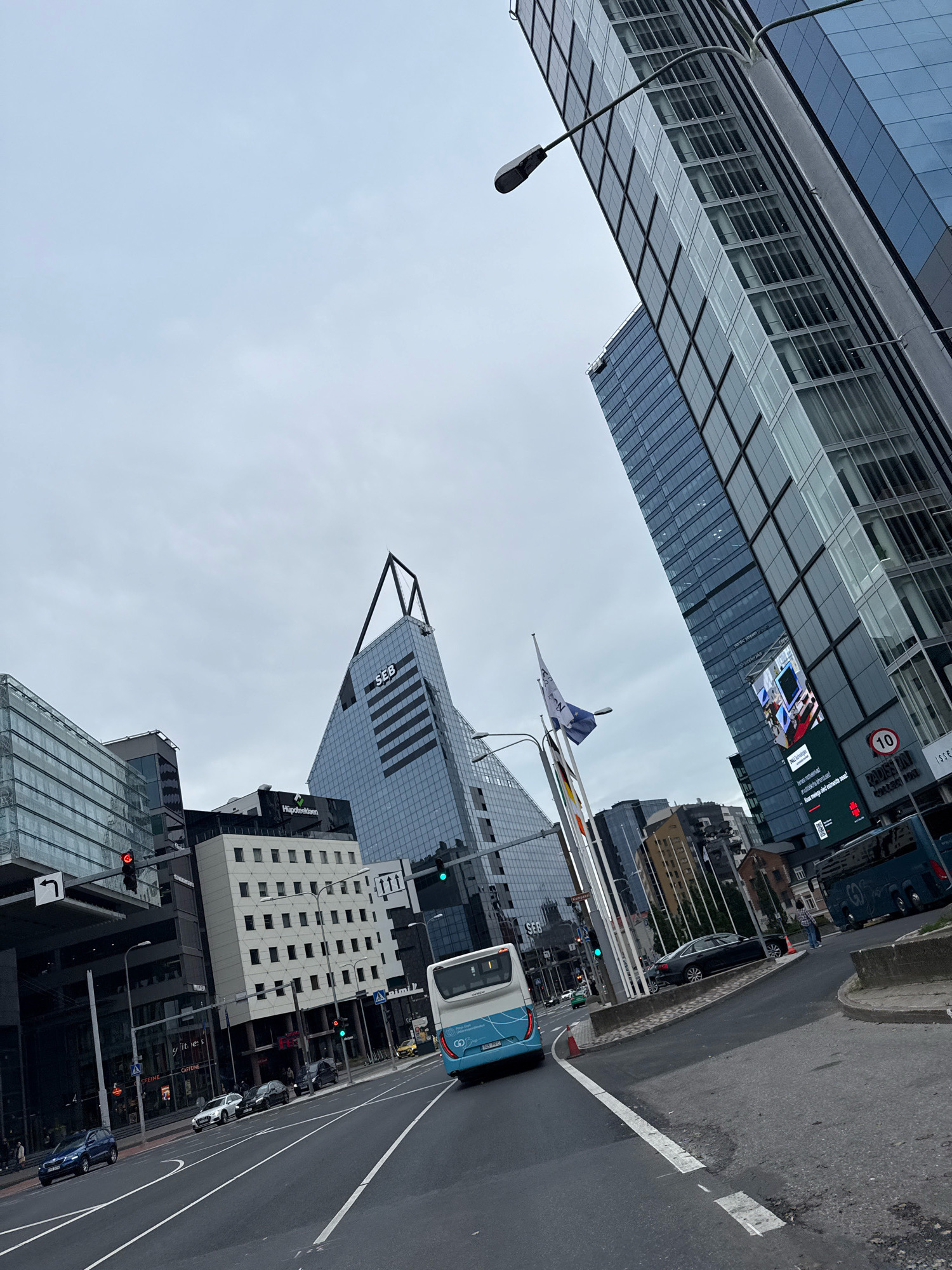 The image captures a city street scene in Tallinn, Estonia, likely in the business district due to the presence of modern skyscrapers. The perspective is low, looking up at the buildings and across the street. A blue and white public transport bus is in the foreground, moving away from the viewer. Several cars are visible on the road, including a blue SUV and a black sedan. The buildings are a mix of modern glass and steel structures and older, lower-rise buildings. One prominent skyscraper has a distinctive pointed roof and the "SEB" logo. Another large glass building on the right has a digital billboard displaying text and images. Traffic lights are visible, and the sky is overcast. The street appears to be a multi-lane road with pedestrian crossings.