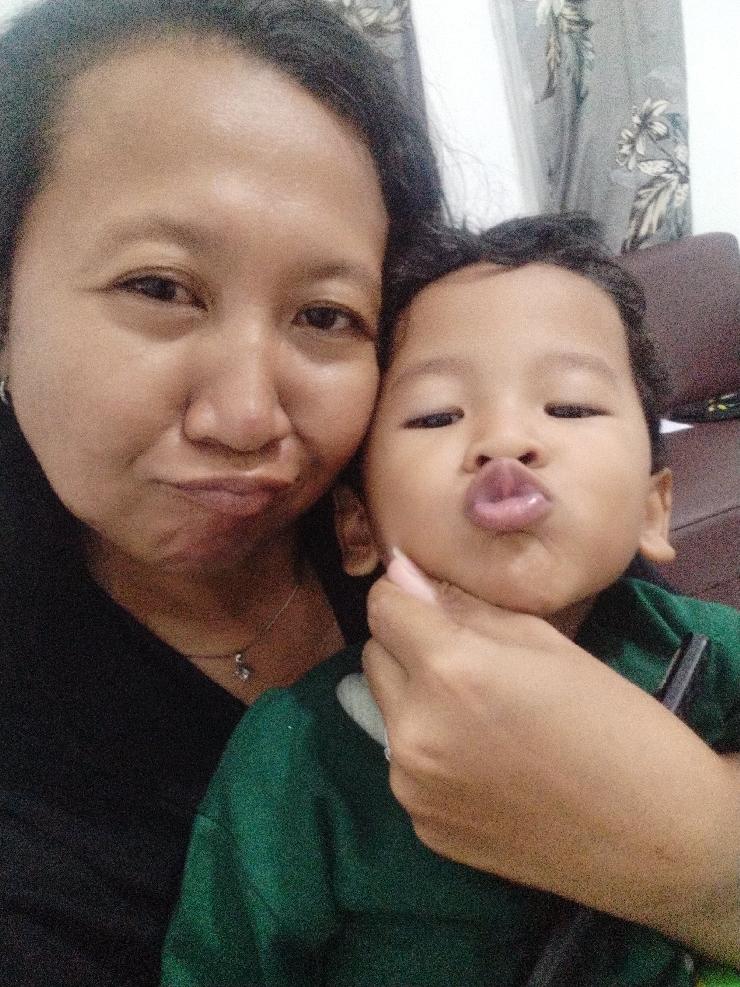 In Karangsentosa, Indonesia, a selfie is captured showing a woman and a young child. The woman, positioned on the left, faces the camera with a slight smile, her head angled towards the child. She has dark hair and is wearing a black top. A silver necklace with a pendant is visible around her neck. The child, on the right, is held by the woman and puckers their lips towards the camera, dressed in a green shirt. The background consists of a patterned curtain and a portion of a dark brown sofa. The lighting suggests an indoor setting.