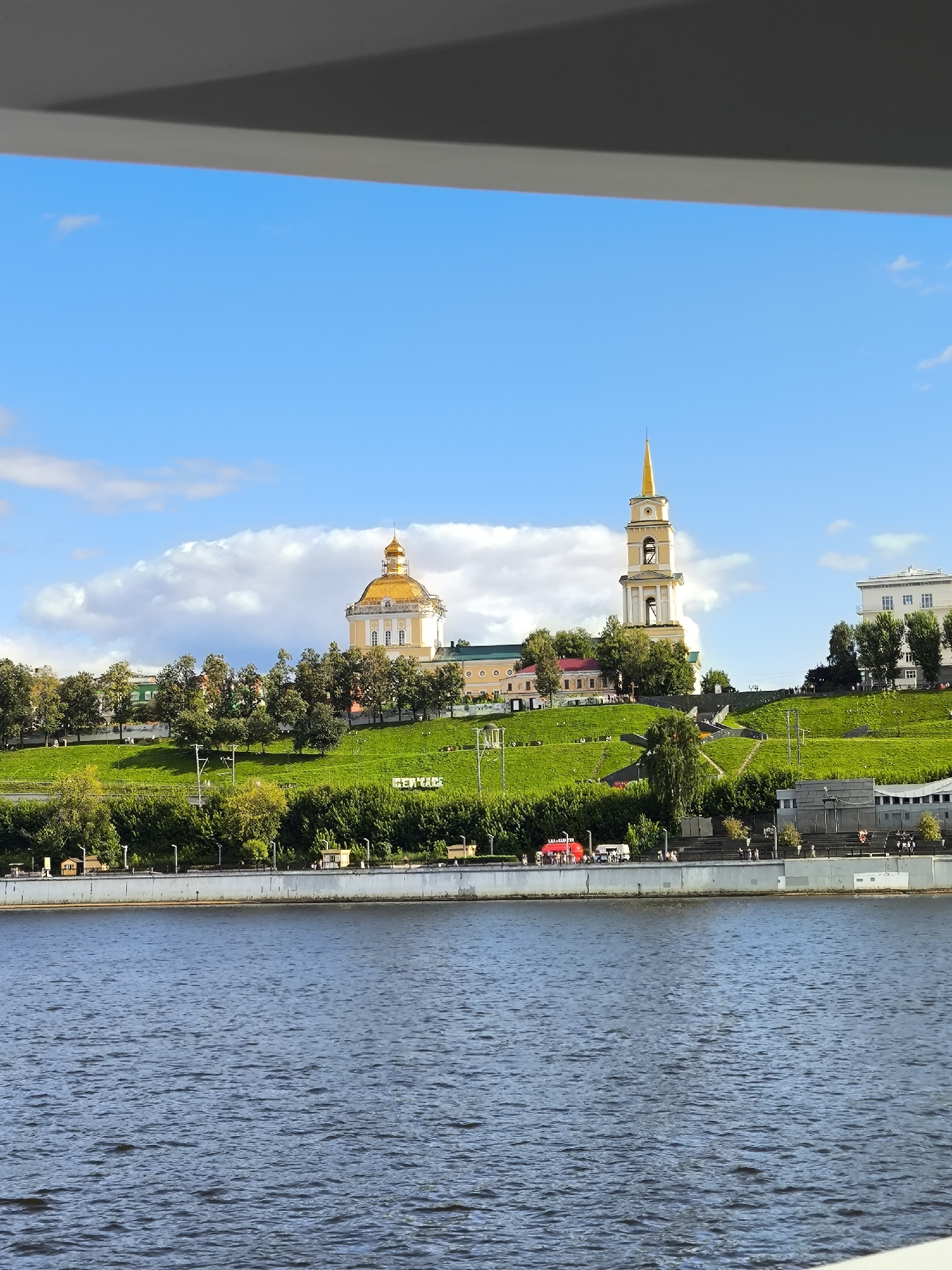 This image captures a waterfront view of the city of Perm, Russia. In the foreground, a wide, deep blue river with gentle ripples dominates the lower portion of the frame. Beyond the river, a steep, grassy embankment rises, dotted with lush green trees and a pathway with stairs leading upwards.
Prominently situated on the embankment are several buildings. A large, cream-colored structure with a golden dome is visible on the left, and to its right, a tall, slender bell tower with a golden spire stands out. Further to the right, a modern, white building with multiple windows is present. On the grassy slope, some people are visible walking along the pathways and near the waterfront. A red vehicle and a white van are also parked along the waterfront promenade. The sky above is a bright blue, with scattered white clouds. The overall scene depicts a serene and picturesque urban landscape.