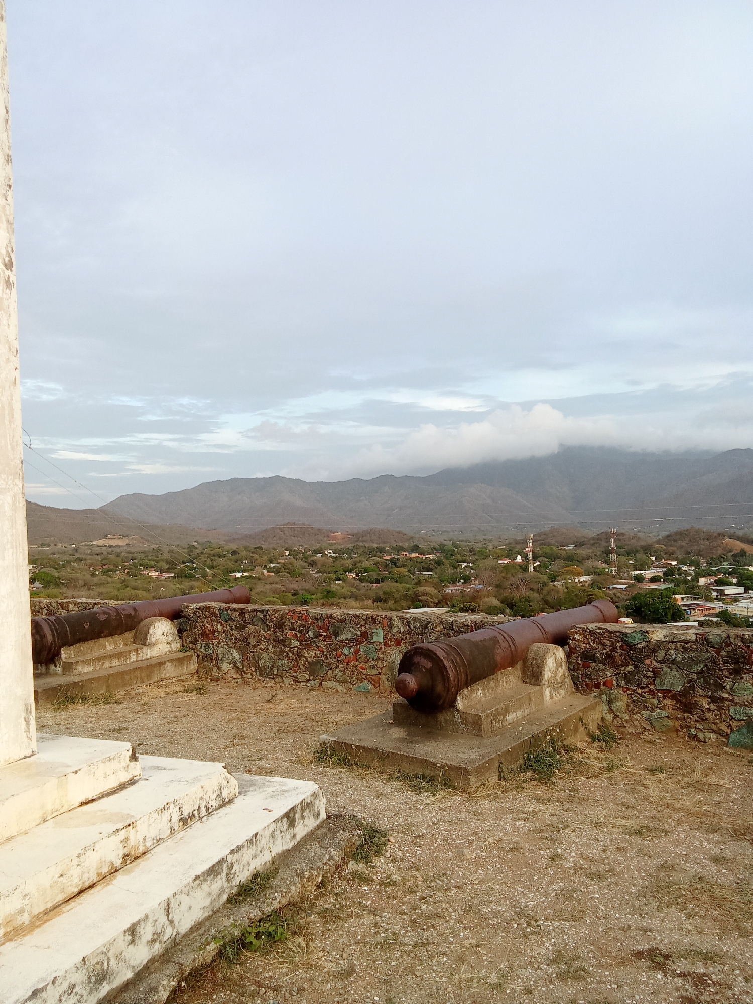 The image shows two cannons positioned on a stone and concrete structure overlooking a town, likely in the city of Juan Griego, Venezuela. The cannons are rusted and mounted on concrete bases, situated along a low wall constructed of stone and mortar. In the background, a town with buildings and trees stretches toward hills and mountains under a cloudy sky. A white architectural structure is partially visible in the foreground to the left of the frame. The ground in the foreground is dry and covered with gravel.