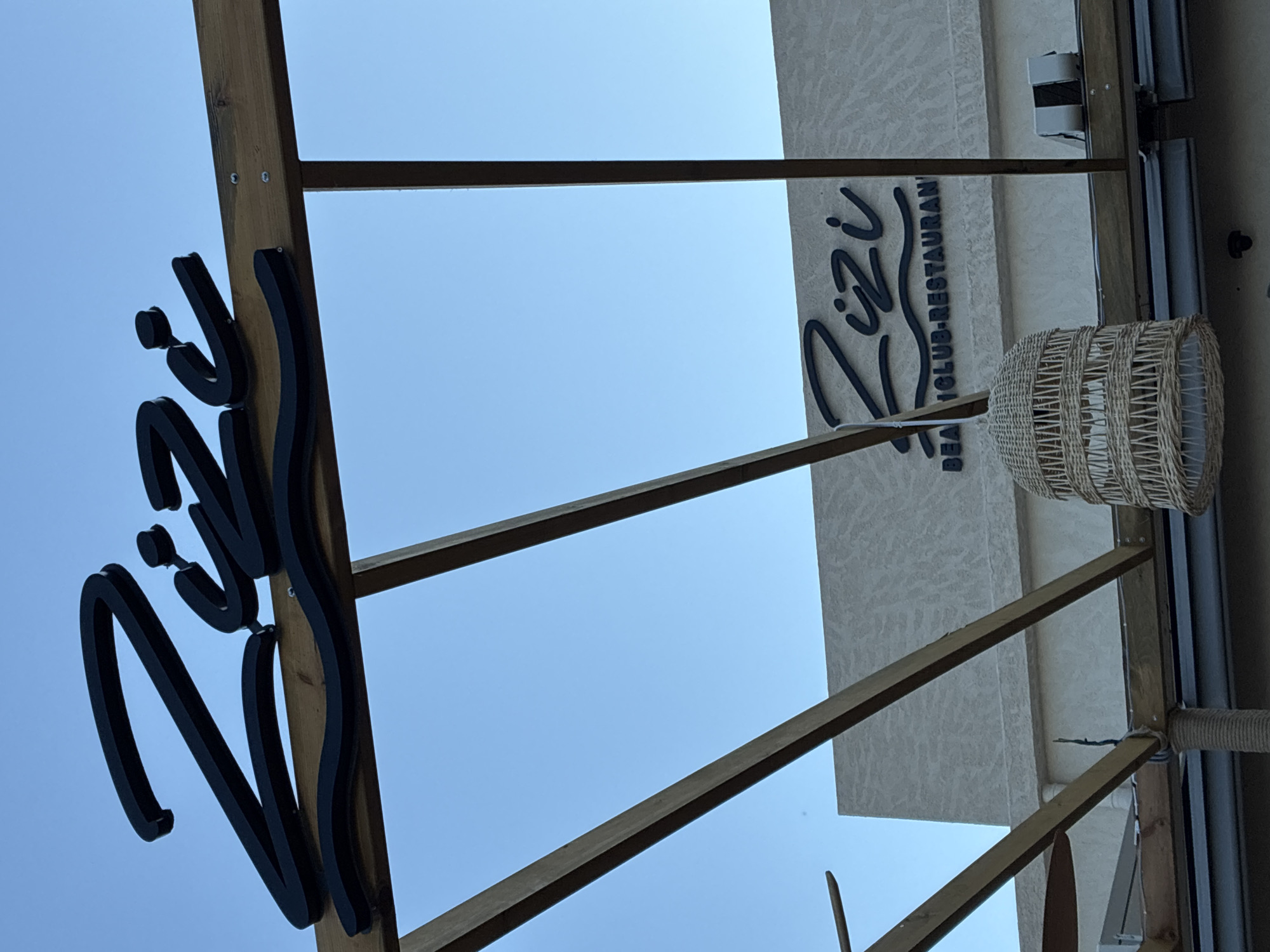 The image shows a close-up view of signage for a beach club and restaurant called "Zizi" in Alanya, Turkey. The signage is in black, stylized script, with the larger "Zizi" logo positioned prominently on a wooden pergola structure against a clear blue sky. Below the main logo, a smaller sign reads "BEACH CLUB-RESTAURANT". A woven lamp shade hangs down from the pergola, adding a decorative element. The building behind the signage is a light-colored structure with textured walls. The overall scene suggests a sunny outdoor establishment, likely a popular spot for dining and leisure.