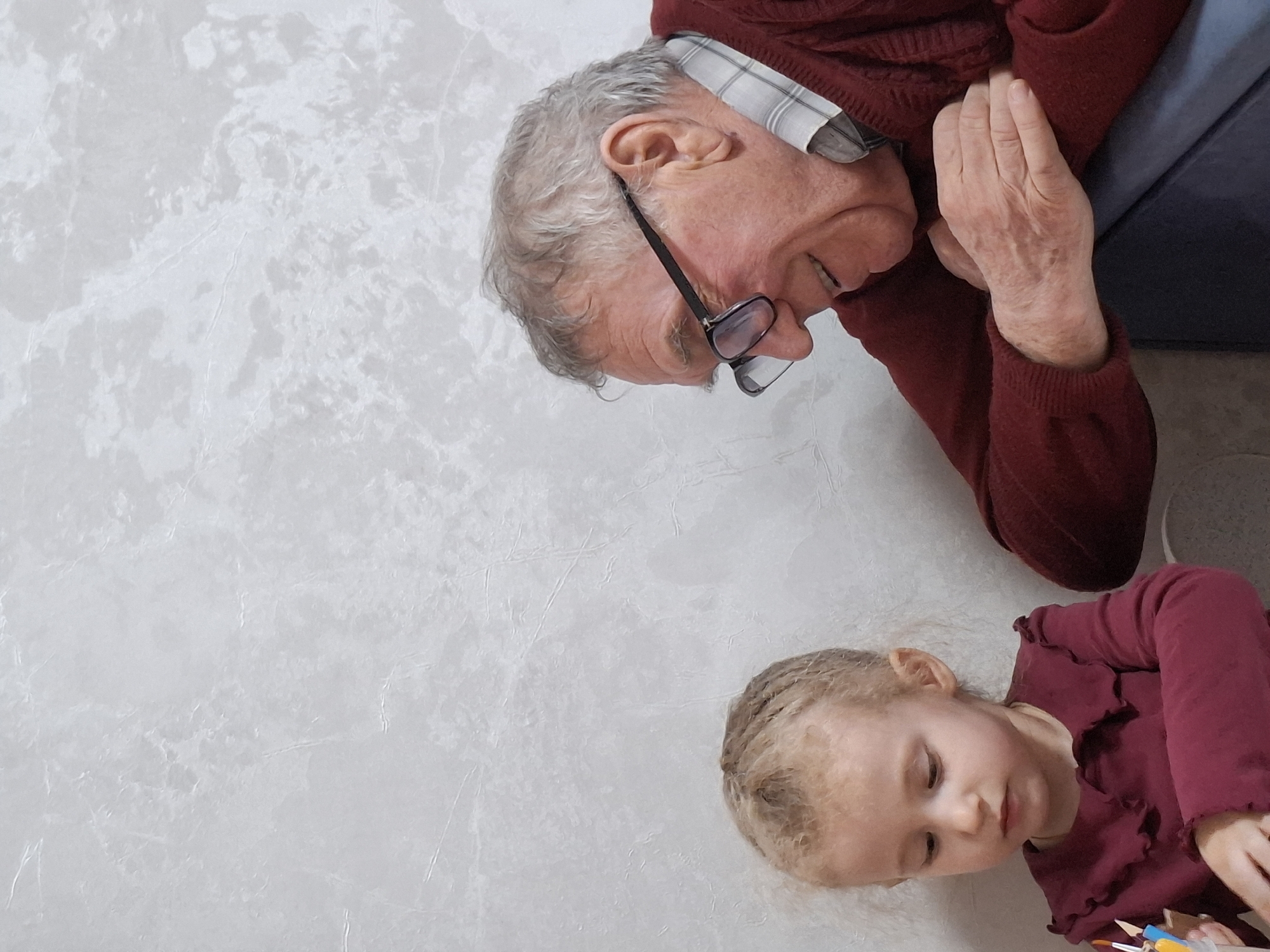 An overhead shot captures an elderly man and a young child, likely a grandfather and granddaughter, sharing a quiet moment indoors in Chaikovsky, Russia. Both are positioned on a light, subtly patterned surface, possibly a floor or large mat. The man, with grey hair and glasses, is wearing a red sweater over a plaid shirt. He is looking down with a gentle, focused expression and a slight smile. His hands are visible, one resting near his chest, the other by his side. Behind him, a dark blue or grey piece of furniture is visible. Below him, to the right of the frame, a young child with blonde hair, styled in braids or small buns, wears a matching red long-sleeved top. The child is also looking down, seemingly engrossed in an activity, with a small collection of art supplies, including colored pencils or brushes, visible near her hands. The scene conveys a warm, intimate interaction, possibly during a creative play session. No visible text is present in the image.