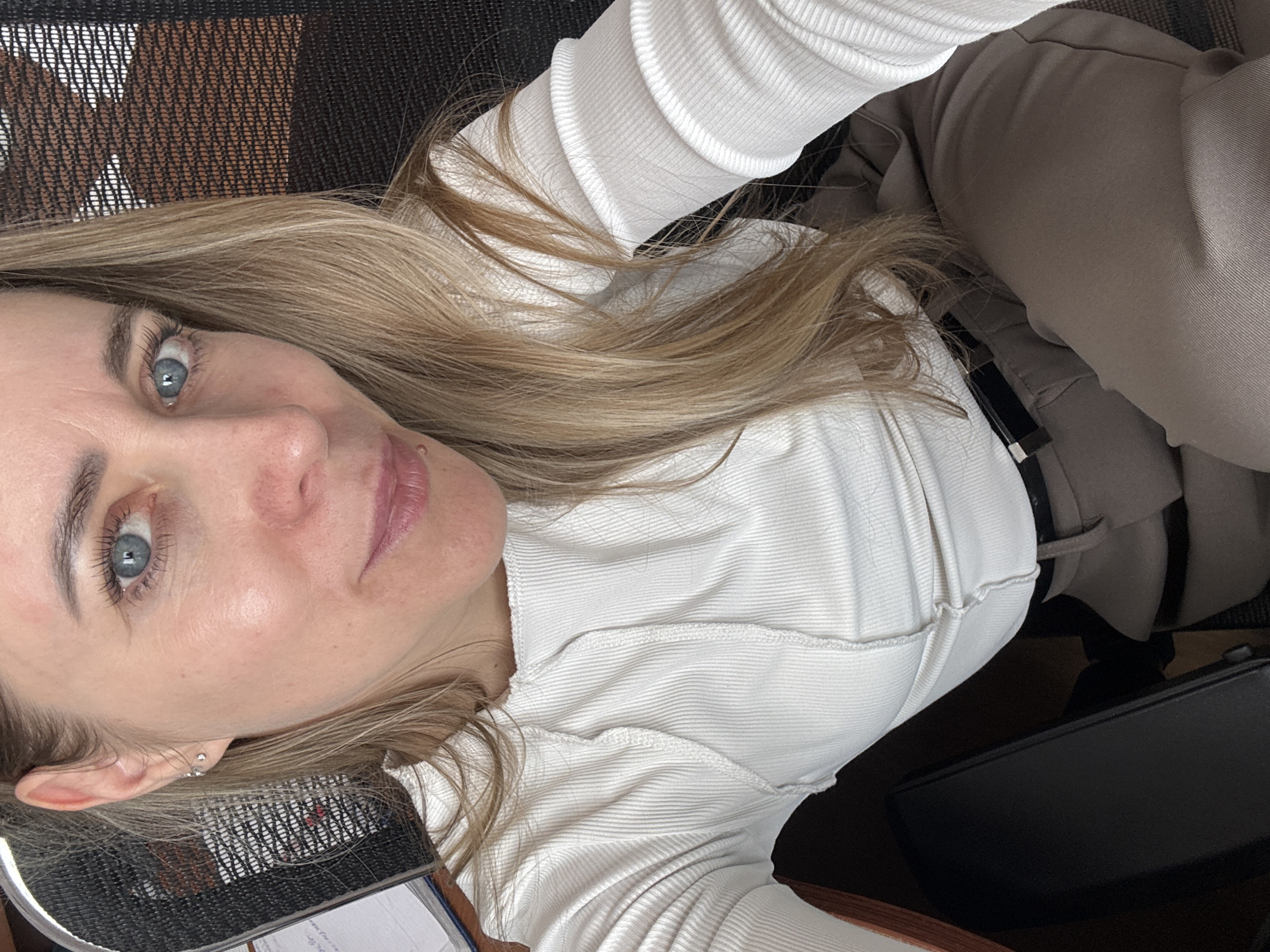 A blonde woman with light blue eyes is looking directly at the camera. She is wearing a white long-sleeved shirt and light brown trousers. The background includes what appears to be an office chair with a woven back and a black object that could be a laptop. There is no information to place the location of this image in Chaikovsky, Russia.