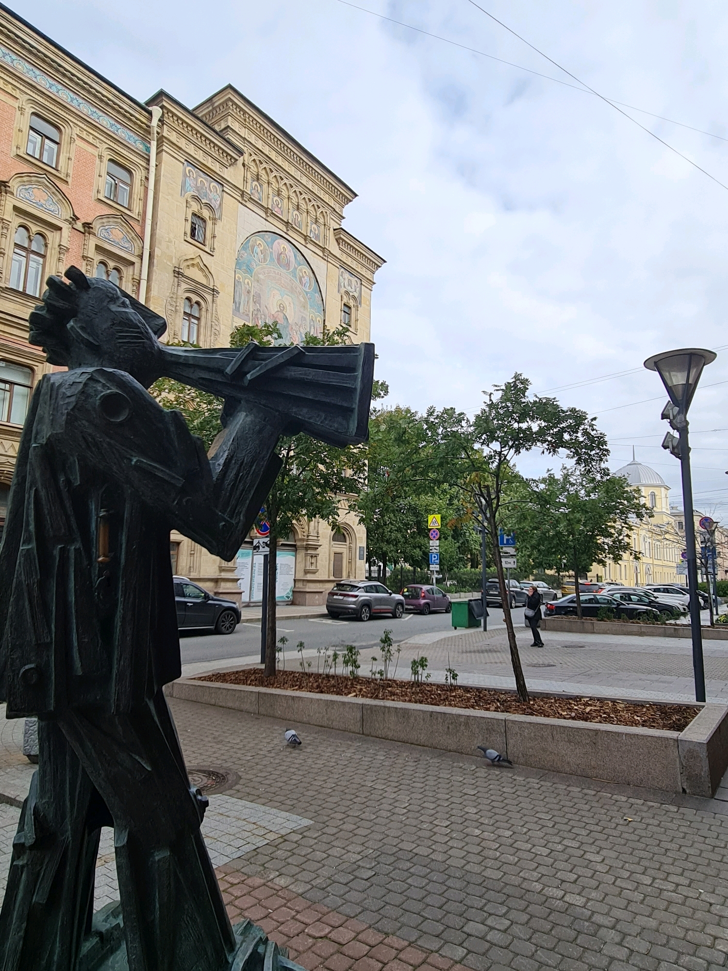 This is a medium shot, eye-level, exterior photograph of a statue of a person playing a horn, located in Saint Petersburg, Russia. The statue is made of dark metal and is positioned in the foreground, to the left of the image. The background features a building with a yellowish facade, ornate architectural details, and a large mural. Across the street, there are parked cars, trees, a street lamp, and a dome-shaped building in the distance. The sky is overcast. The ground is paved with cobblestones. A pigeon is visible on the cobblestones in the middle ground. The statue appears to be of a man in a cape blowing into a large, rectangular horn. The building behind the statue has multiple windows and decorative elements along its upper facade, including a large arched mural depicting figures. A street is visible with cars driving and parked along its side. A street lamp with multiple light fixtures stands on the right side of the image.