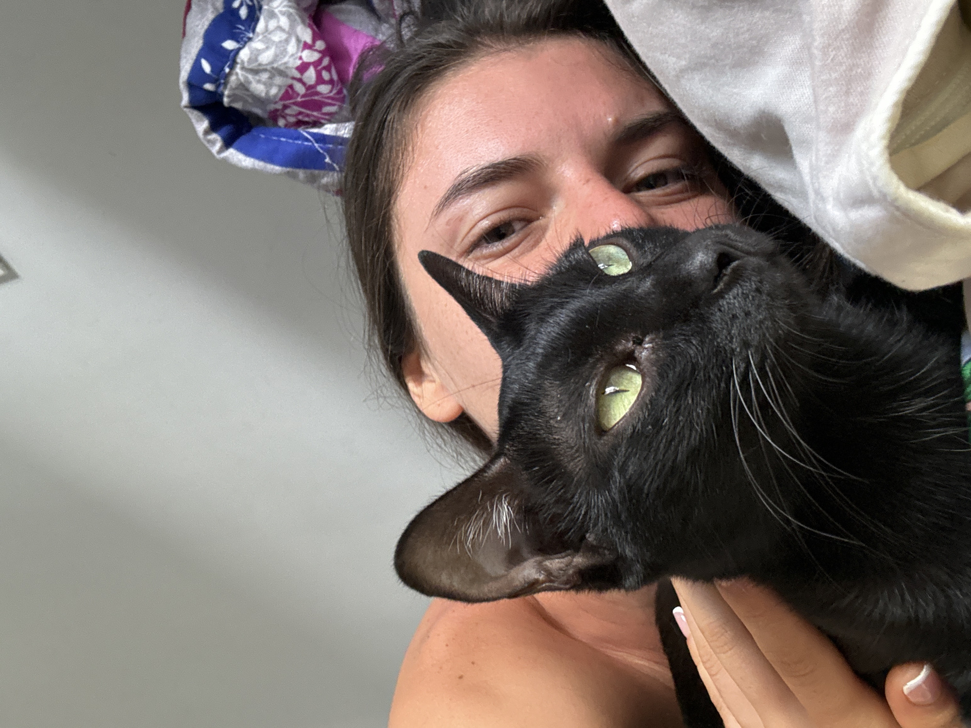 A person is holding a black cat close to their face. The person has light skin, dark hair, and is looking at the camera. The cat has green eyes and a sleek coat. A colorful fabric with floral and geometric patterns is visible above the person's head, and a white fabric is to the right of their head. The background is a plain white or light-colored wall. No location information is discernible from the image.