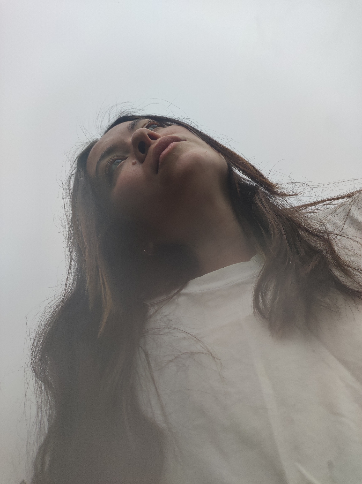 A low-angle photograph shows a young woman with long brown hair looking upwards, presumably towards the sky, which is overcast. She wears a white t-shirt. Her facial features are visible, including her eyes, nose, and mouth. The city of Фоки, Russia is not discernible in the photograph.