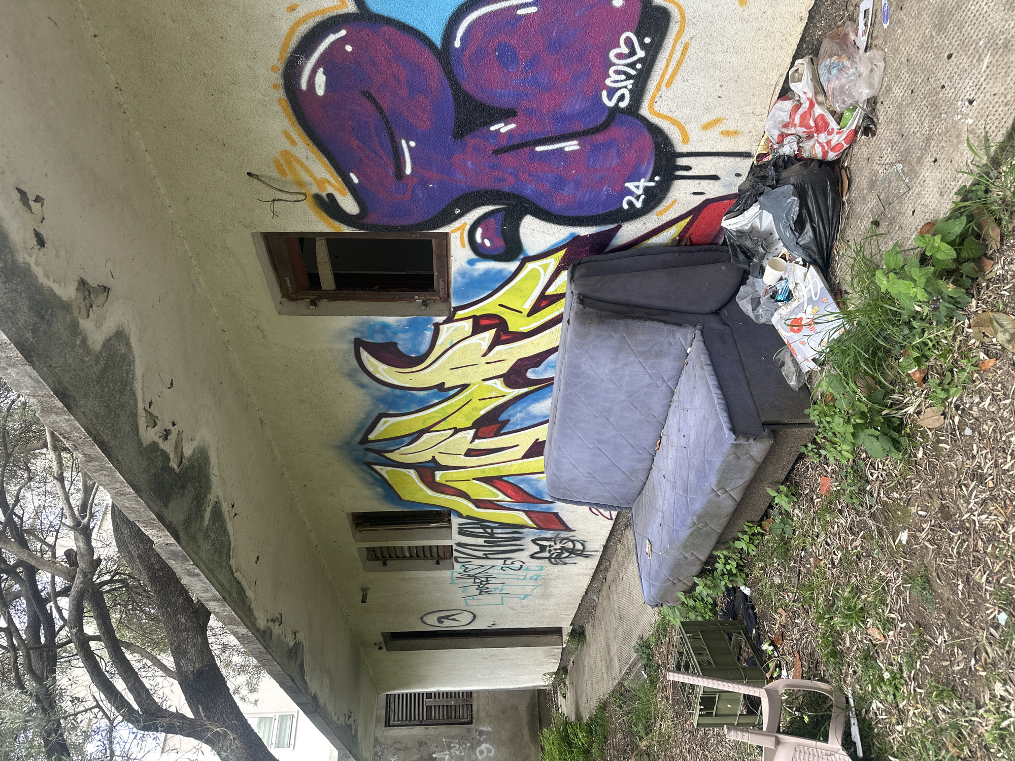 The image depicts an exterior scene of an abandoned or neglected building. The building is concrete, covered in graffiti, including purple, yellow, and blue tags. Several window openings are visible, some boarded up or missing frames. A discarded gray sofa sits against the wall, partially obscured by garbage bags and assorted trash. Weeds and grass grow around the base of the building, and a plastic chair lies on the ground nearby. A tree with bare branches is on the left side of the frame. The location is likely in Budva, Montenegro, based on context clues. There are no people visible in the image. The scene suggests urban decay and neglect.