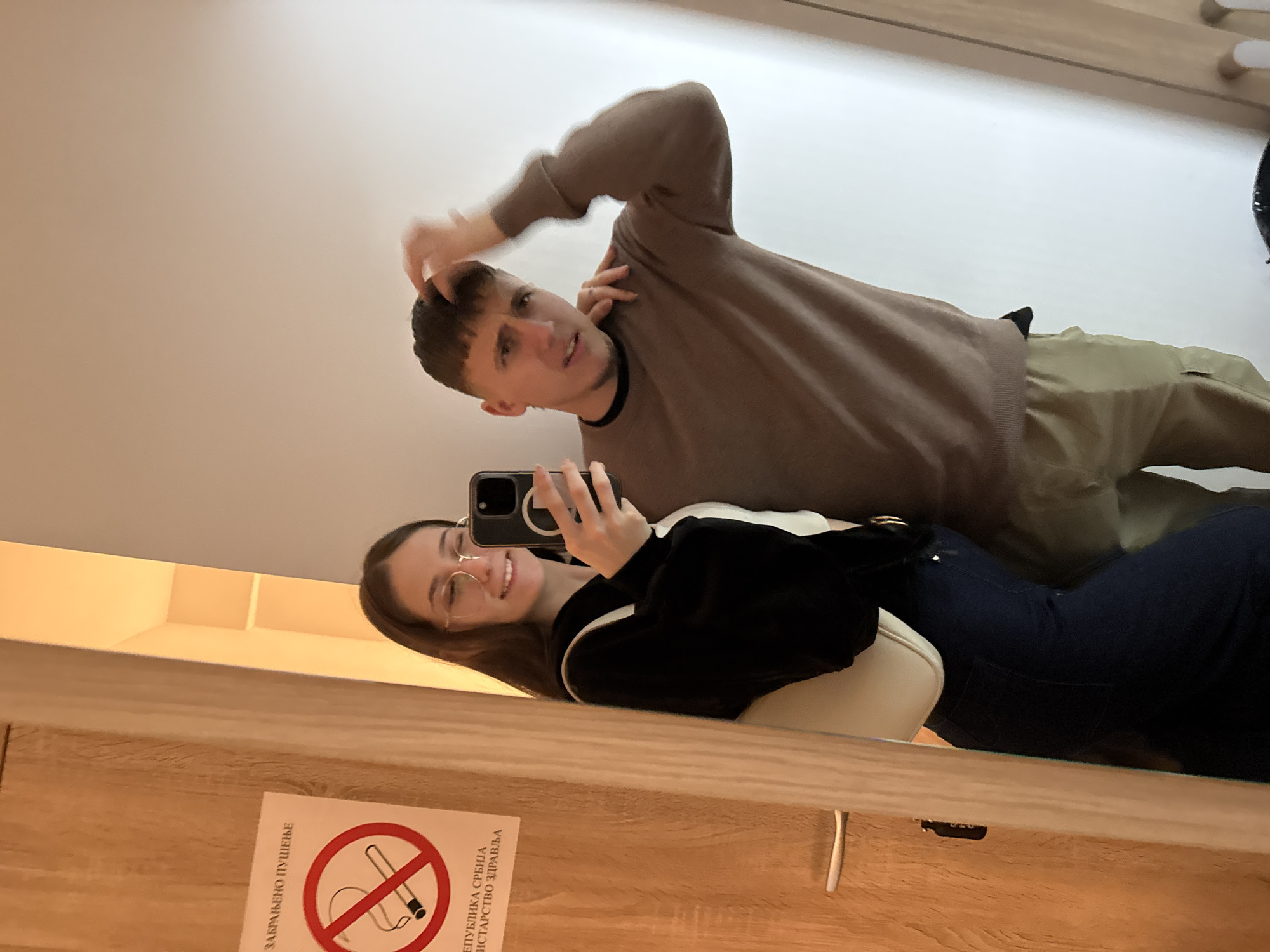 A mirror selfie captures two individuals indoors within the city of Vozdovac Urban Municipality, Serbia. The location is indicated by a visible "ЗАБРАЊЕНО ПУШЕЊЕ" (No Smoking) sign with "РЕПУБЛИКА СРБИЈА" (Republic of Serbia) text. The female subject, possessing long dark hair and eyeglasses, stands on the left, holding a dark smartphone in her right hand to take the photograph, smiling. She is dressed in a black top over a white layer, dark blue jeans, and carries a light-colored shoulder bag. Her left hand rests on the right shoulder of the male subject. The male subject, with short dark hair, is positioned on the right, wearing a brown sweater over a black t-shirt and light-colored cargo pants. He looks towards the camera with an open mouth and a somewhat perplexed facial expression, his right hand situated on the back of his head. A wooden door with a silver handle is partially visible on the far left, adjacent to the no-smoking sign. The background on the right consists of a plain, light-colored wall.