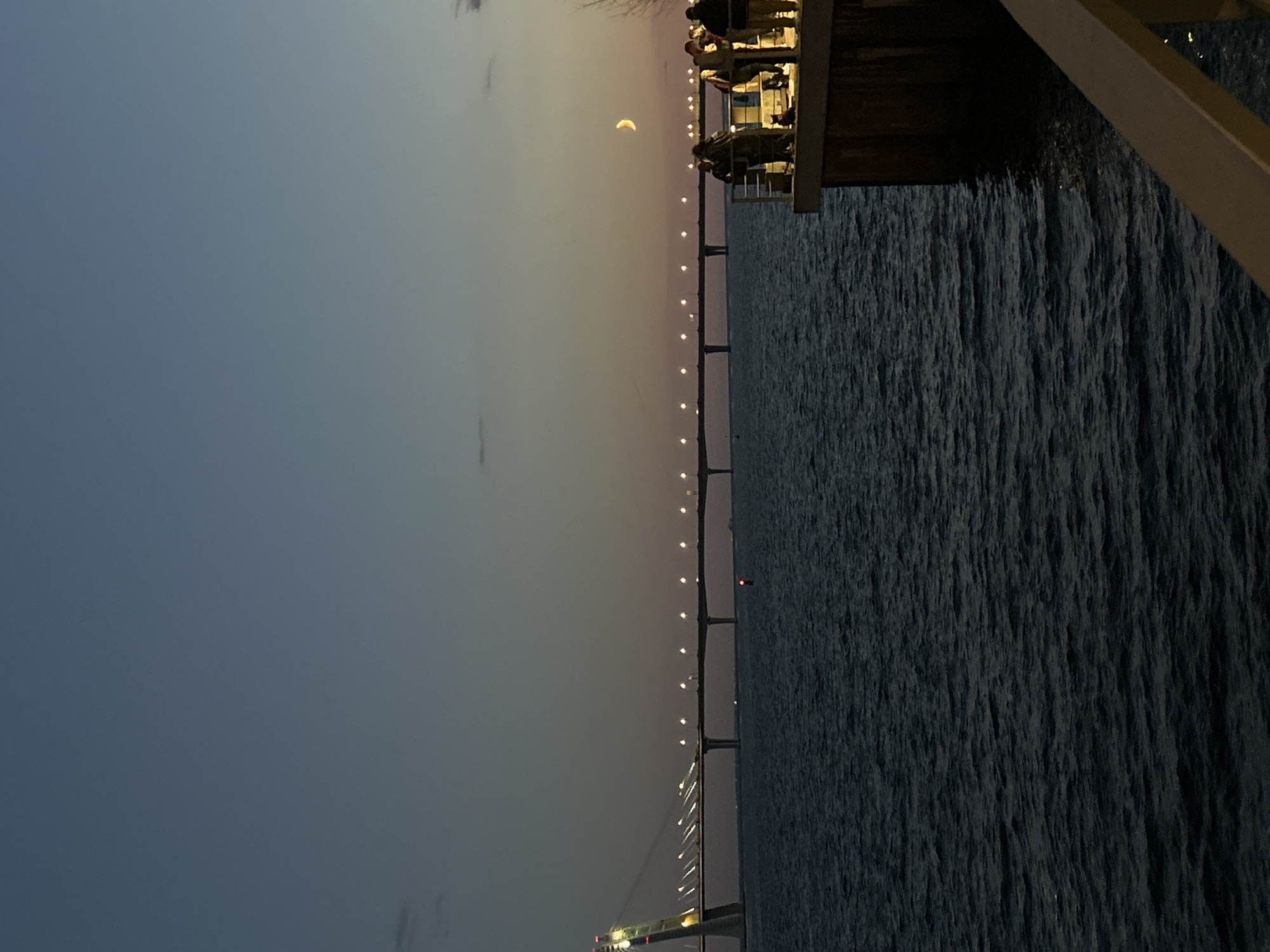 At dusk, a long, illuminated bridge spans across a dark body of water. A faint crescent moon is visible in the twilight sky. On the right side of the image, a group of people are standing on a pier, silhouetted against the dim light. The water's surface is choppy, with small waves. The scene is likely of the Western High-Speed Diameter in Saint Petersburg, Russia, a modern toll road that crosses the Neva Bay.