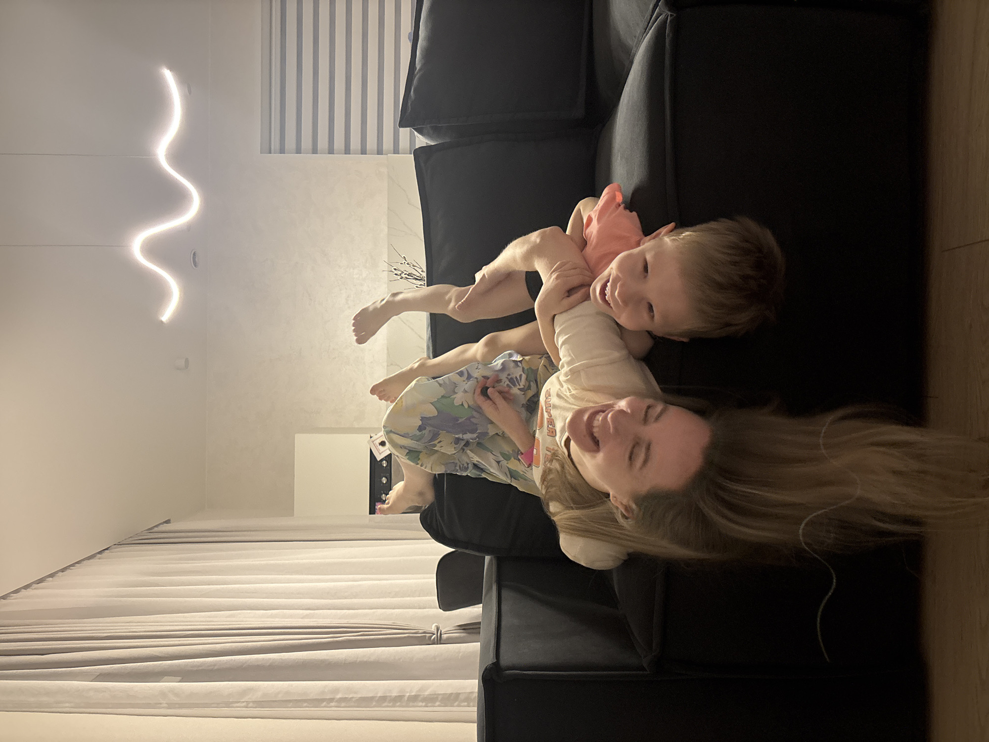 A photograph taken indoors shows a woman and a child lying on a dark-colored sofa. The woman is wearing a light-colored t-shirt and a floral dress. The child is wearing a pink top and dark shorts. Both are smiling. The room has curtains on the left and a wavy light fixture on the ceiling. The sofa is set against a wall with striped wallpaper. The location is assumed to be in an apartment in Chaikovsky, Russia.