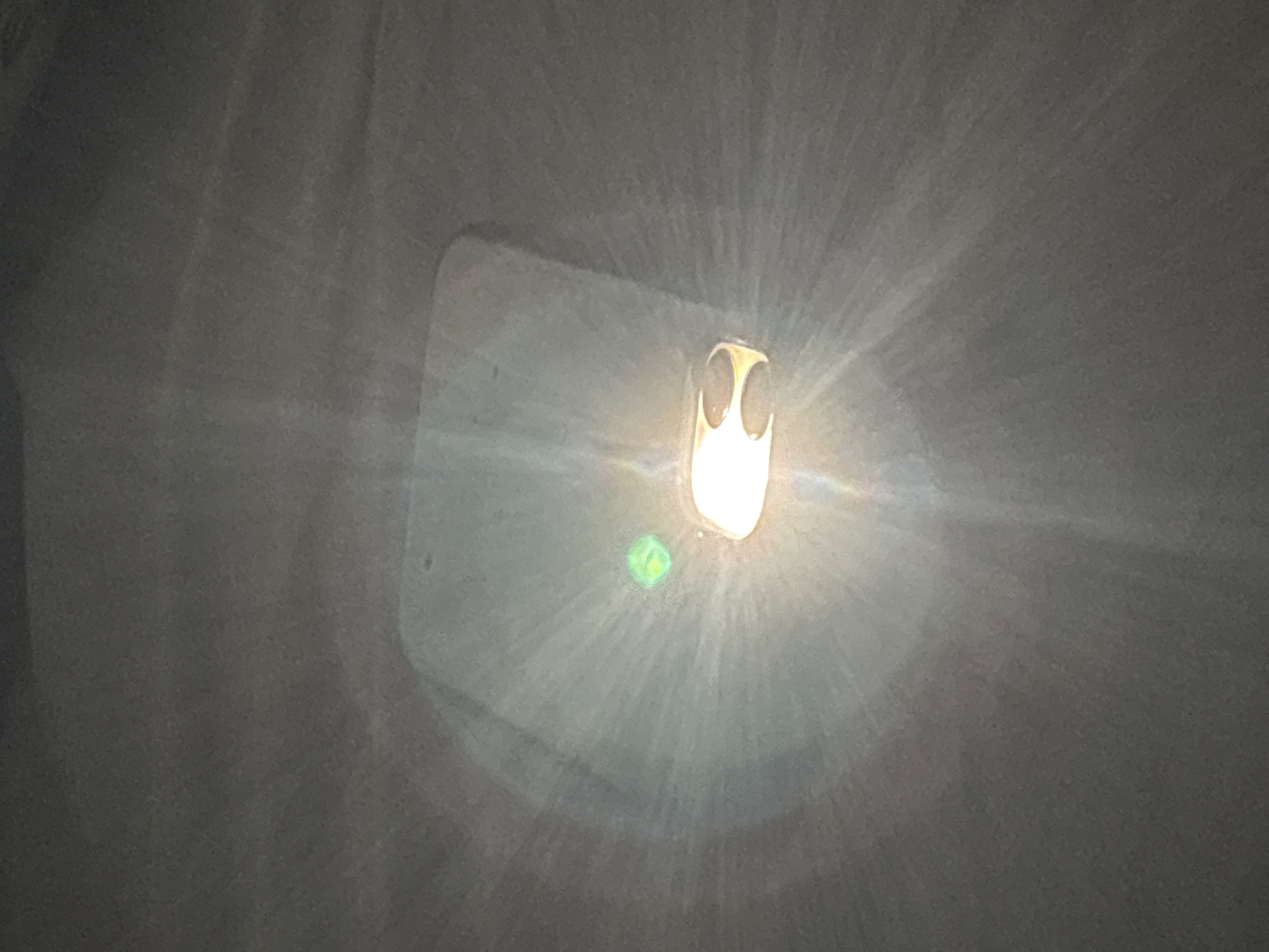 The image displays an overhead perspective of an interior space, presented as located in Angwan Sayawa, Nigeria. A prominent, intensely bright light source is positioned centrally, emitting strong white-yellow light rays that create a starburst lens flare effect across the frame. This main light fixture features a light-colored, possibly metallic, housing with an irregular shape and a distinct decorative cutout on its right side. To the immediate left of the primary light, a smaller, square-shaped, vibrant green light source is visible. The dominant background is a uniform, medium-grey, textured surface, consistent with a ceiling or upper wall, which exhibits subtle lighter variations. No individuals, additional distinct objects, or specific actions are discernible within the frame.