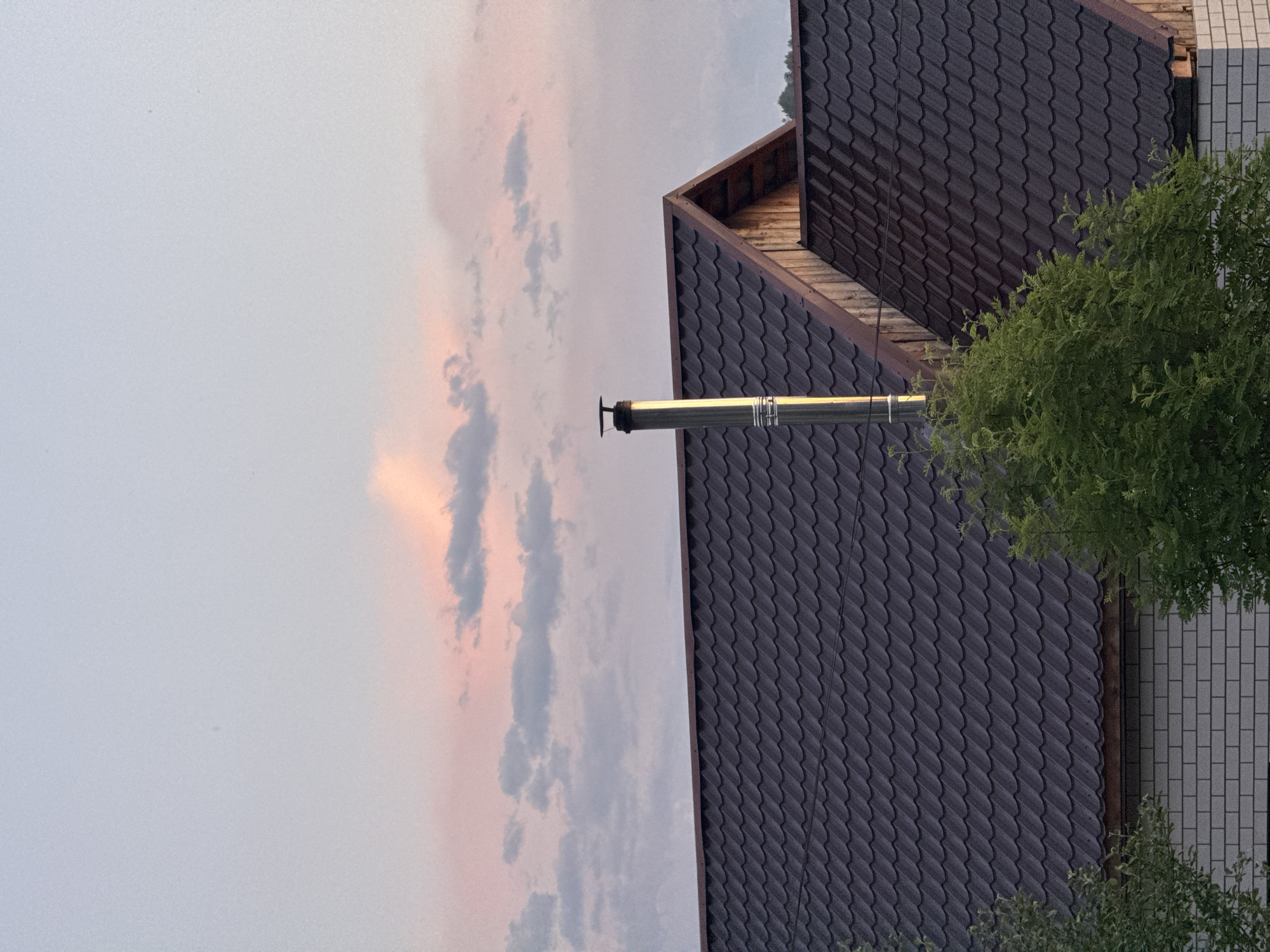 The image depicts a residential building's roof and sky, likely in the town of Soloneshnoye, Russia, given the context. The sky above is filled with pale pink and blue clouds, suggesting it's either sunrise or sunset. A silver metal chimney stands tall on the dark, wavy tiled roof of a house. The roofline of another section of the building, made of wooden planks, is visible to the right. In the foreground, lush green trees partially obscure the view of the white brick facade of the house.