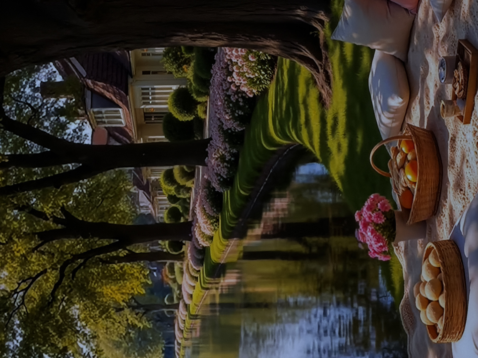 This image captures a tranquil outdoor scene, likely set in Sofiivska Borshchahivka, Ukraine, showcasing a blend of natural beauty and elegant landscaping.
In the immediate foreground, a cozy picnic is arranged on a light-colored, patterned blanket spread on lush green grass. Several soft pillows, including white and light pink ones, are placed for comfort. A picnic basket filled with oranges or similar fruits is visible, alongside another basket brimming with bread rolls. A small wooden tray holds a cup and other items, while a vase containing vibrant pink cluster flowers, possibly hydrangeas, adds a touch of color.
The picnic setup is situated beside a calm canal or river, whose surface reflects the surrounding trees and the soft sky. Along the far bank of the waterway, a meticulously landscaped garden unfolds, featuring rows of neatly shaped green bushes, reminiscent of topiary, and an extensive stretch of flowering plants bearing delicate pink and white blooms. Large, verdant trees with strong, dark trunks and branches frame the scene, their foliage rich green.
In the background, an elegant, grand building, possibly a manor or stately home, is visible. It features a distinctive reddish-brown roof, multiple windows with white frames, and classic architectural details. The lighting suggests a warm late afternoon or early evening, with long shadows stretching across the grass, contributing to a serene and idyllic atmosphere. No visible text is present in the image.