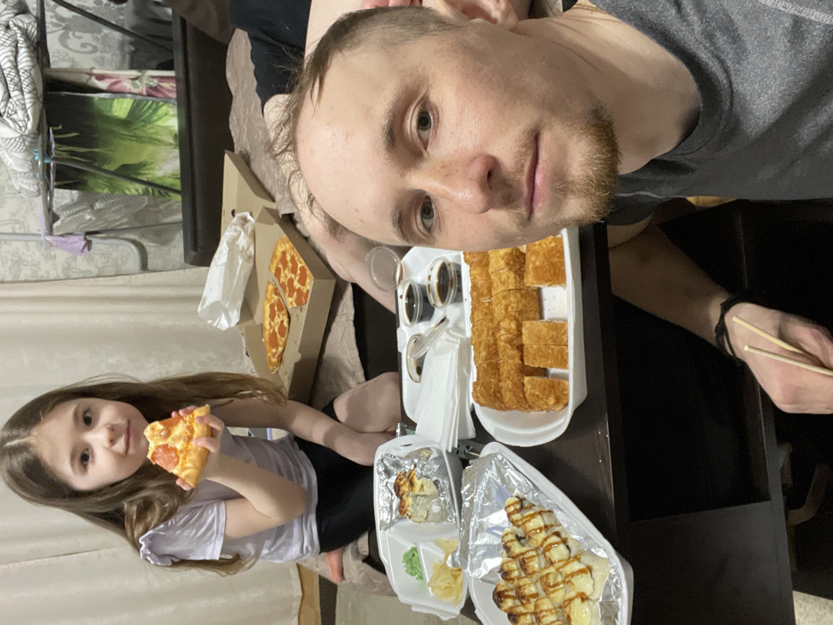 The image depicts an indoor domestic setting in the city of Лубянки, Russia, focusing on a shared meal involving at least three individuals. In the foreground, an adult male with short brown hair, a light beard, and a grey t-shirt is taking a selfie, holding a pair of wooden chopsticks in his right hand. To his left, a young female child, estimated to be 5-8 years old, with long brown hair and wearing a light purple top and dark bottoms, is seated, holding a single slice of pepperoni pizza in her right hand, looking directly at the camera.
A dark brown table or low surface between the individuals and the camera displays multiple food items and containers. These include: an open cardboard pizza box containing two slices of pepperoni pizza; a white rectangular tray holding approximately 18-20 pieces of golden-brown, breaded or fried sushi rolls, some cut into large rectangular blocks and others into smaller squares; a second white rectangular tray containing several pieces of baked or gratinated sushi rolls with a creamy topping, arranged on aluminum foil; and a smaller white multi-compartment container with green wasabi paste, yellow pickled ginger, and a single baked sushi roll. Additionally, several small transparent plastic cups containing dark liquid (consistent with soy sauce) and white plastic utensils are present.
In the mid-background, partially obscured by the food and foreground individuals, the lower legs of a third individual are visible, resting on a sofa or bed covered with a brown or tan textured blanket. The background wall features light-colored patterned wallpaper, with a light-colored curtain visible on the left. In the upper right background, a portable clothes drying rack is visible, holding dark green and purple fabric items. The scene is illuminated by artificial indoor lighting.