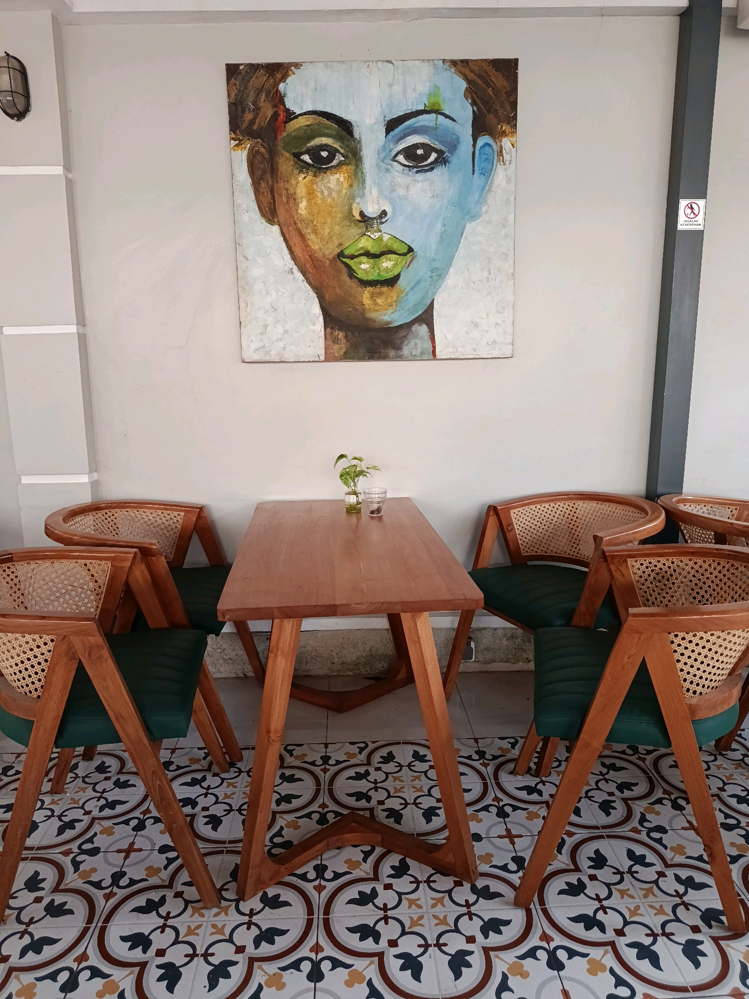 The image shows an outdoor seating area at a cafe, likely in Dawe, Indonesia. A long wooden table is set up with six wooden chairs featuring cane backs and green upholstered seats. The chairs are arranged around the table, with three on each side. On the table, there is a small glass vase with a green plant and a small glass.
The walls are painted a light grey, and a large, abstract painting of a face with bold colors dominates the wall behind the table. To the right of the painting, there is a black vertical beam with a small sign that reads "JAGALAH KEBERSIHAN," meaning "Keep Clean" in Indonesian, accompanied by a no-smoking symbol.
The floor is tiled with a decorative pattern of interlocking shapes in shades of white, brown, yellow, and dark blue. The overall ambiance suggests a relaxed and stylish cafe environment. The lighting appears to be natural, coming from above and possibly from an unseen open area. There are no people visible in the image.