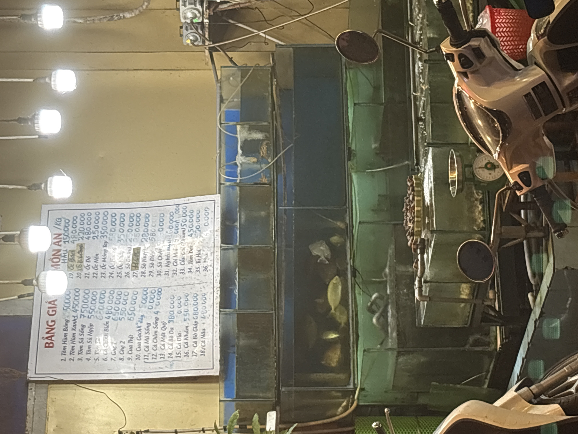 The image shows a price list board outside a seafood restaurant, likely in Nha Trang Ward, Vietnam. The board displays prices for various seafood items like shrimp, crab, and fish. In the background, there are several aquariums filled with live seafood and fish. The lighting suggests it's evening or nighttime. A scooter is parked in the foreground, indicating a street-side establishment. The overall scene depicts a bustling local market or restaurant environment.