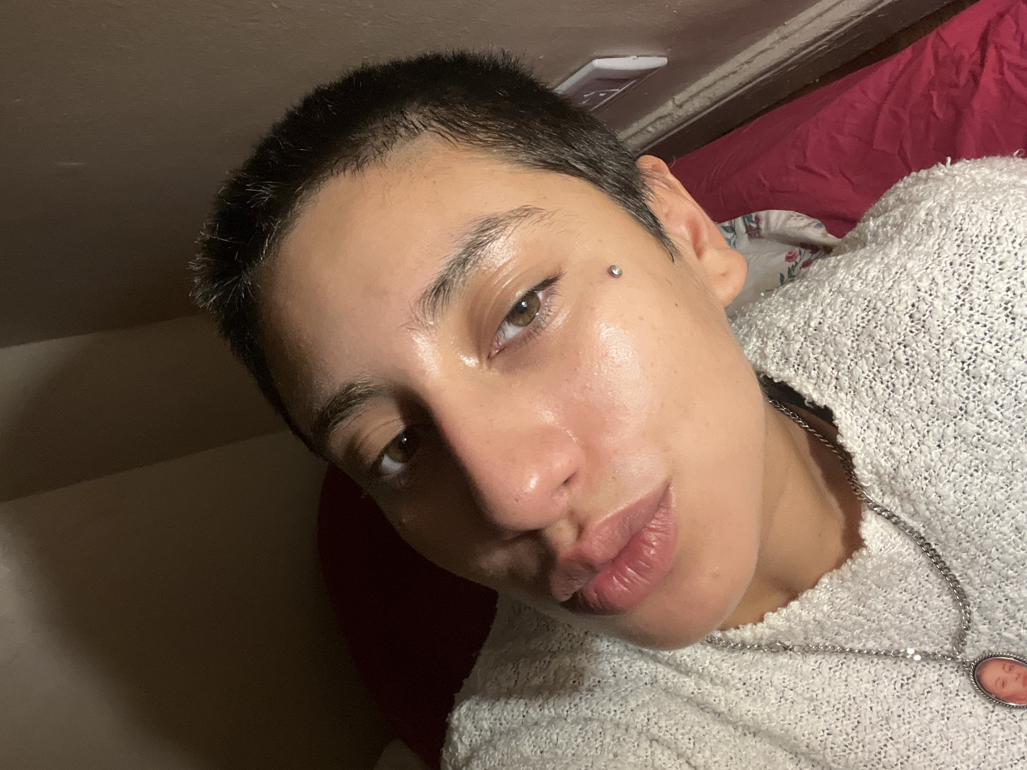 A close-up, selfie-style image depicts a person with short, dark hair and brown eyes. The person has fair skin and a piercing above their left eyebrow. They are wearing a white, textured sweater and a silver necklace with a pendant. The background includes a glimpse of a burgundy sheet and a white object on the ceiling. The environment appears to be an indoor bedroom setting. There is no information that indicates the location in the city of Buenos Aires, Argentina.