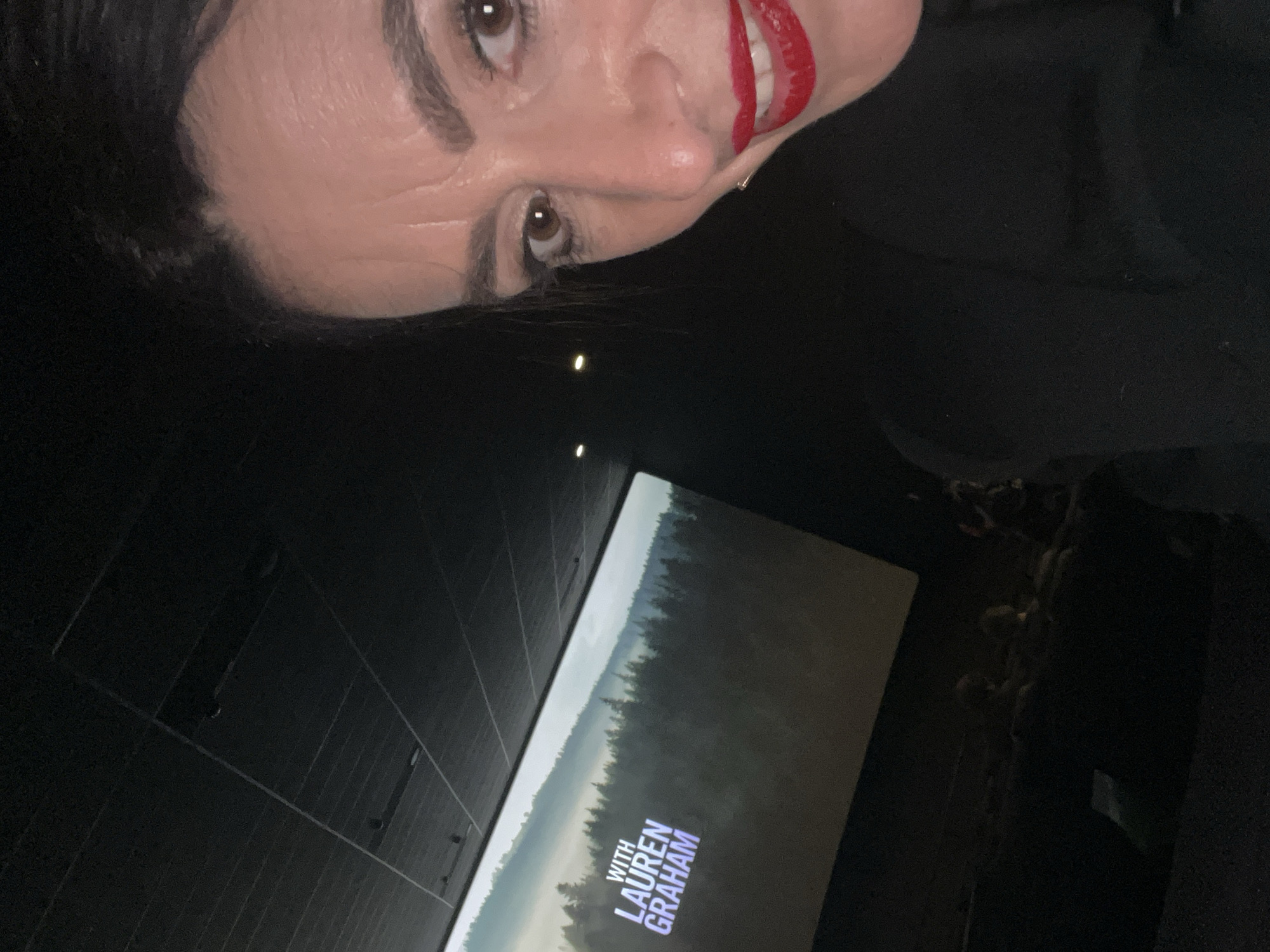 The image is a selfie taken by a woman with dark hair and bright red lipstick. She is looking at the camera with a slight smile. The background appears to be a movie theater or similar venue, given the large screen showing the words "WITH LAUREN GRAHAM" over an image of a forest. The room is dark, but there are faint rows of what appears to be an audience visible in the far right. The surrounding walls are dark and textured with horizontal lines. It is not possible to definitively determine the location as Luxembourg City, Luxembourg, from the image alone.