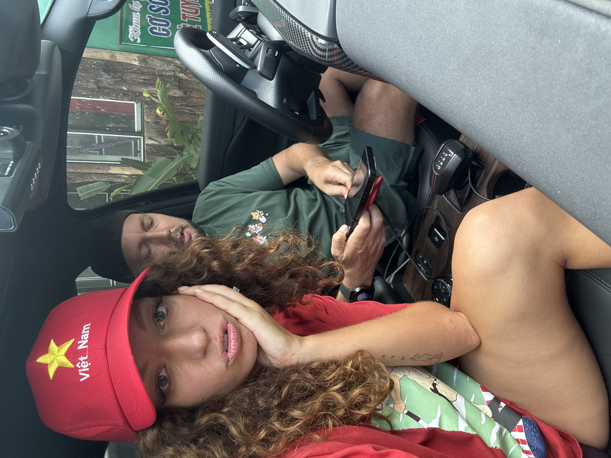Two people are in a car. A woman wearing a red cap with the words "Việt Nam" and a yellow star on the front, and a red shirt, is in the foreground. She has curly hair and is looking at the camera. Behind her is a man wearing a black beanie and a green shirt, looking at a cell phone. The car appears to be parked, with the steering wheel and gear shift visible. Outside the car window is foliage and a building. The location is Suối Giàng, Vietnam.