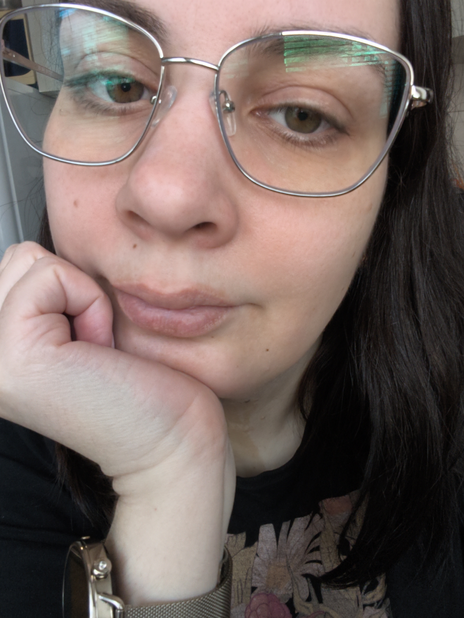 A close-up, chest-up portrait features an individual, likely female, with fair skin, dark hair, and brown eyes, looking towards the viewer. They are wearing silver-framed glasses that have a slightly rectangular or cat-eye shape, with greenish-blue reflections visible on the lenses.
The person's left hand is raised, supporting their chin, with their knuckles gently pressed against their cheek. Their lips are closed in a soft, slightly pouting expression. Dark, straight hair falls over their shoulders, with a section visible on the right side of the frame. Several small moles or freckles are scattered across their face.
The subject is wearing a dark-colored top, possibly a t-shirt, which has a floral pattern featuring muted shades of pink, yellow, and green visible on the chest area. A silver-toned wristwatch with a mesh band is worn on their left wrist.
The image appears to be taken indoors, indicated by the soft lighting and reflections on the glasses, possibly from a screen or window. The background is out of focus, drawing attention to the subject. The overall impression is one of calmness or contemplation. No visible text is present in the image.